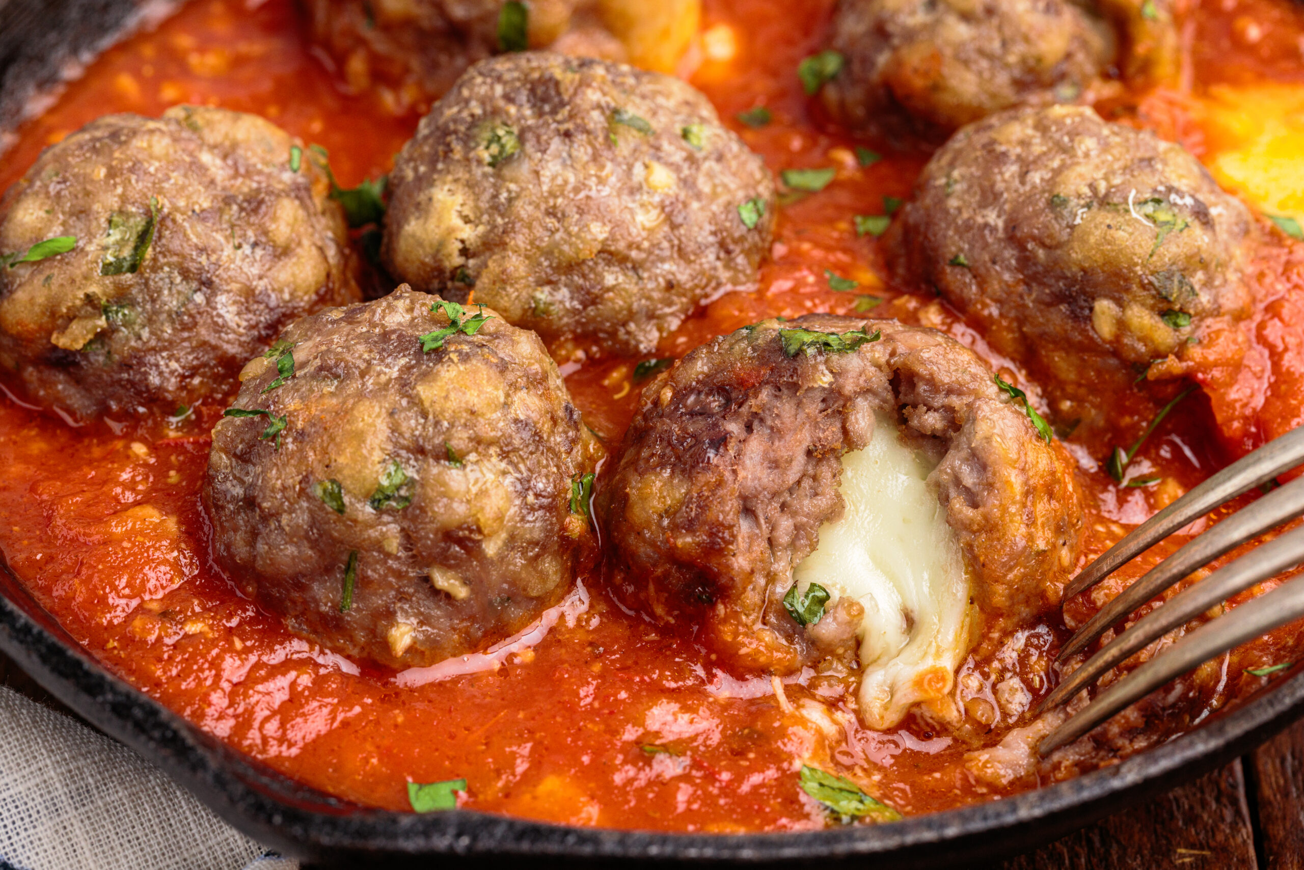 Up close view of Mozzarella Stuffed Meatballs in a cast iron skillet with one meatball cut open by a silver fork.