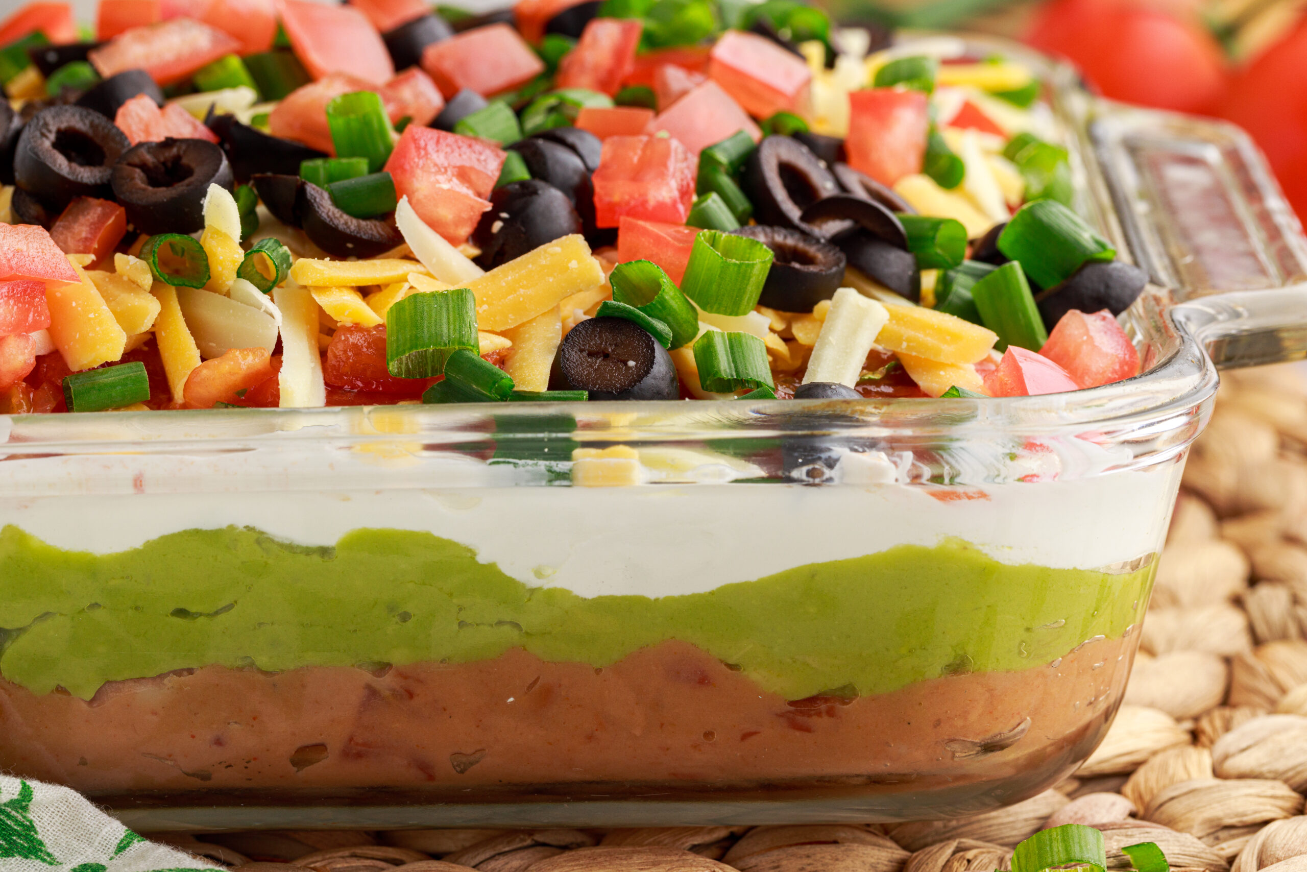 side view of 7-Layer Dip in a clear baking dish, topped with diced tomatoes , black olives, cheese and chives- showing the layers
