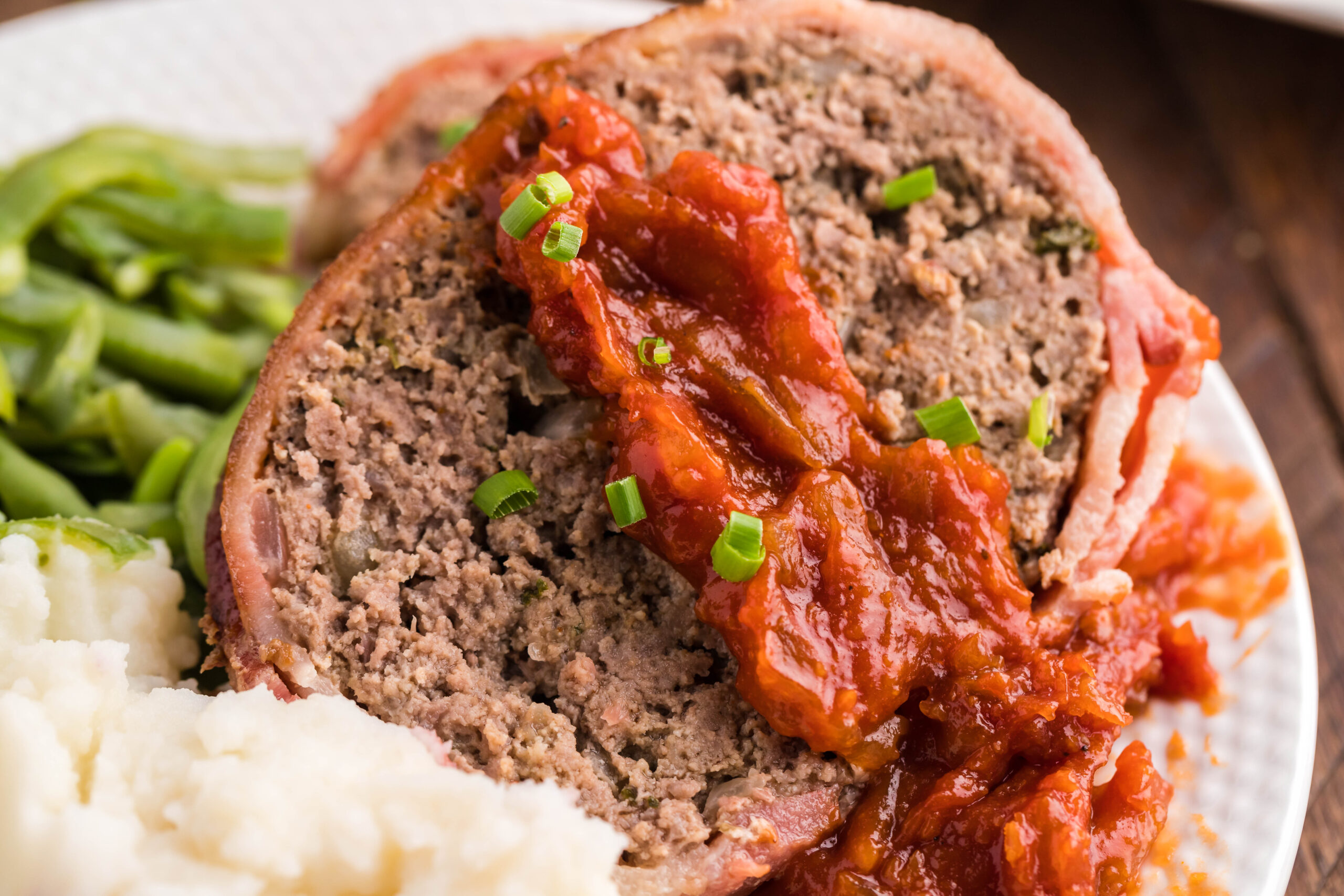 Close up view of a slice of Bacon Wrapped Meatloaf on a white dish served with mashed potatoes and green beans.