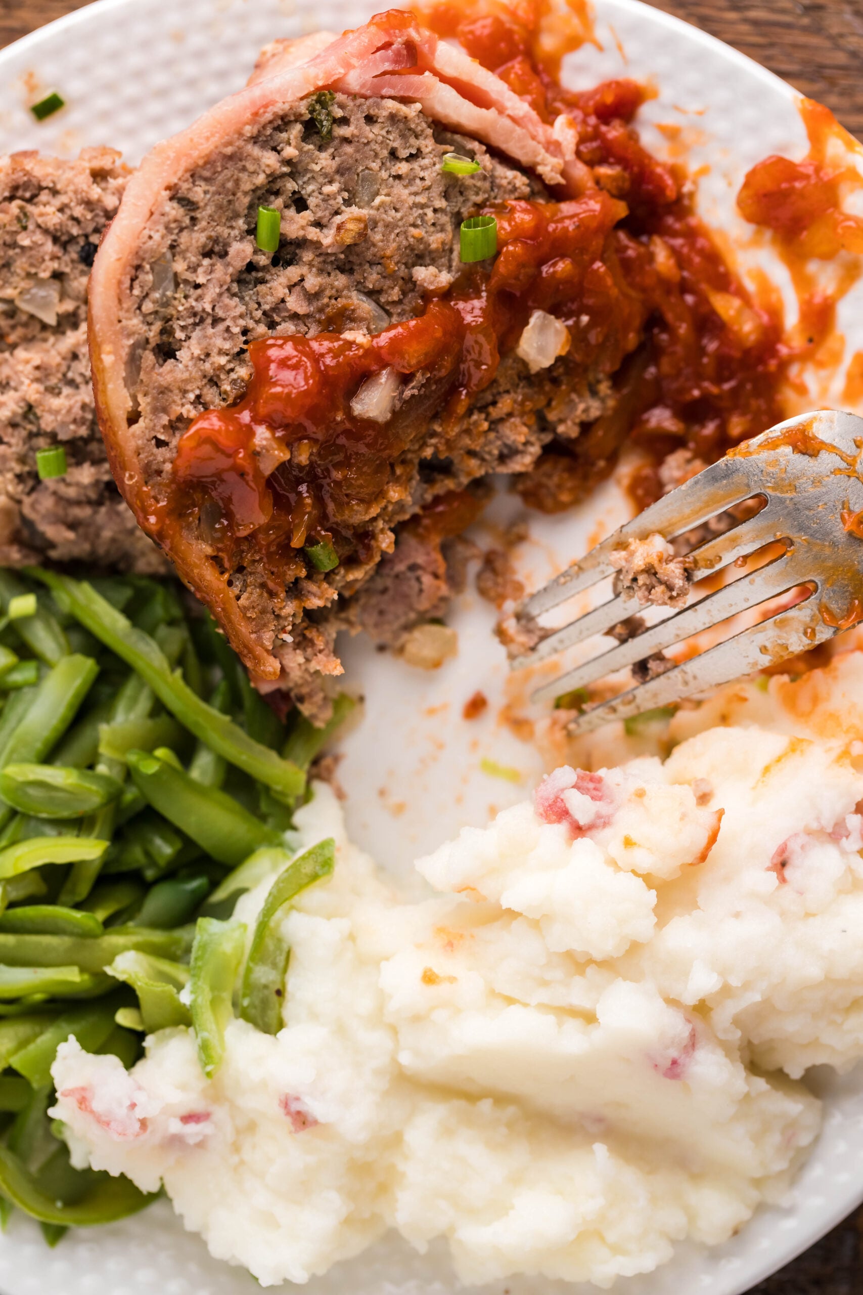 Close up view of a slice of Bacon Wrapped Meatloaf, cut in half by a silver fork, on a white dish served with mashed potatoes and green beans.