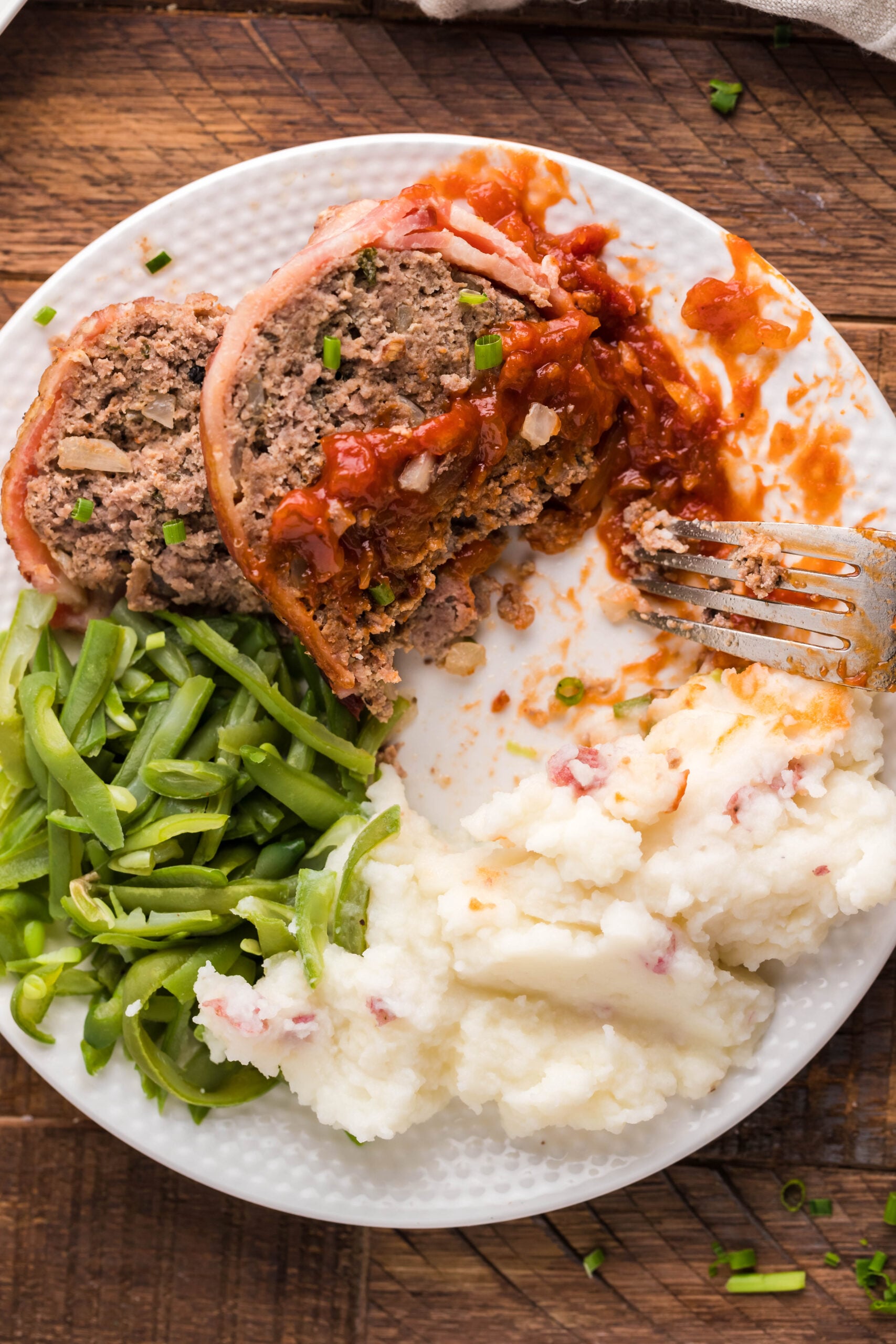 Overhead view of a slice of Bacon Wrapped Meatloaf, cut in half by a silver fork, on a white dish served with mashed potatoes and green beans.