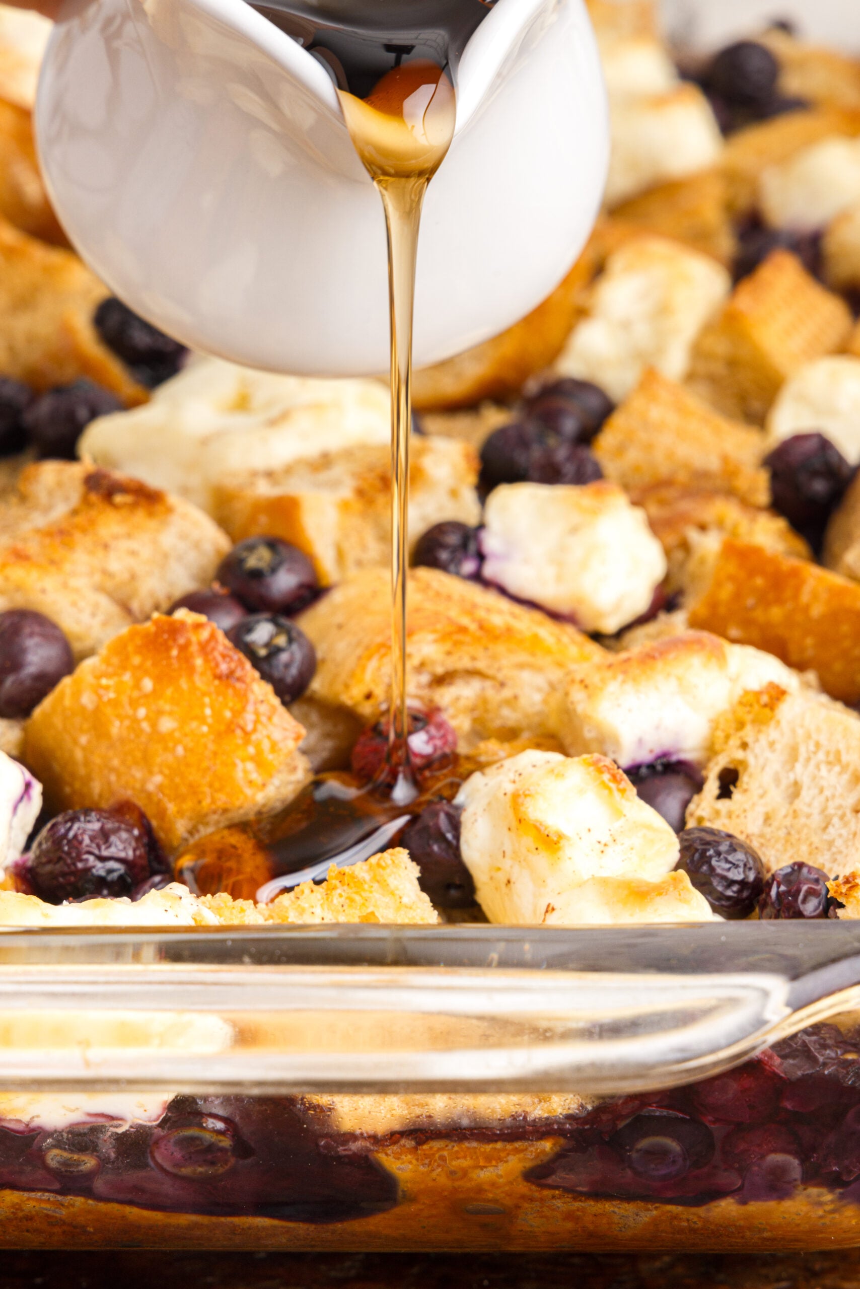 Up close view of Blueberry French Toast Casserole in a clear baking dish with syrup being drizzled over top.