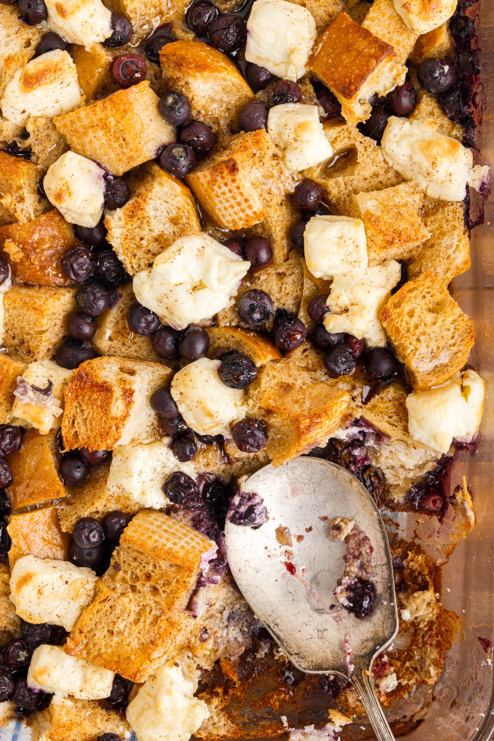 Overhead view of Blueberry French Toast Casserole in a clear dish with a serving removed by a silver serving spoon.