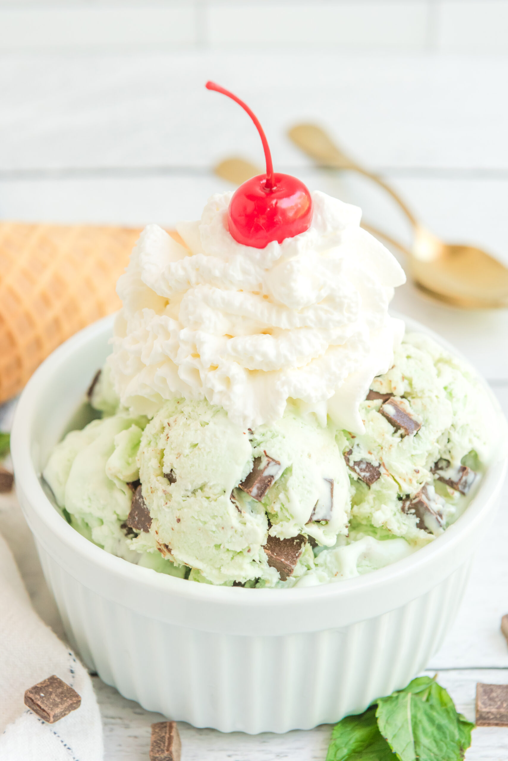 Close up view of scoops of Mint Chocolate Chip Ice Cream in a white bowl garnished with whipped cream and cherry.