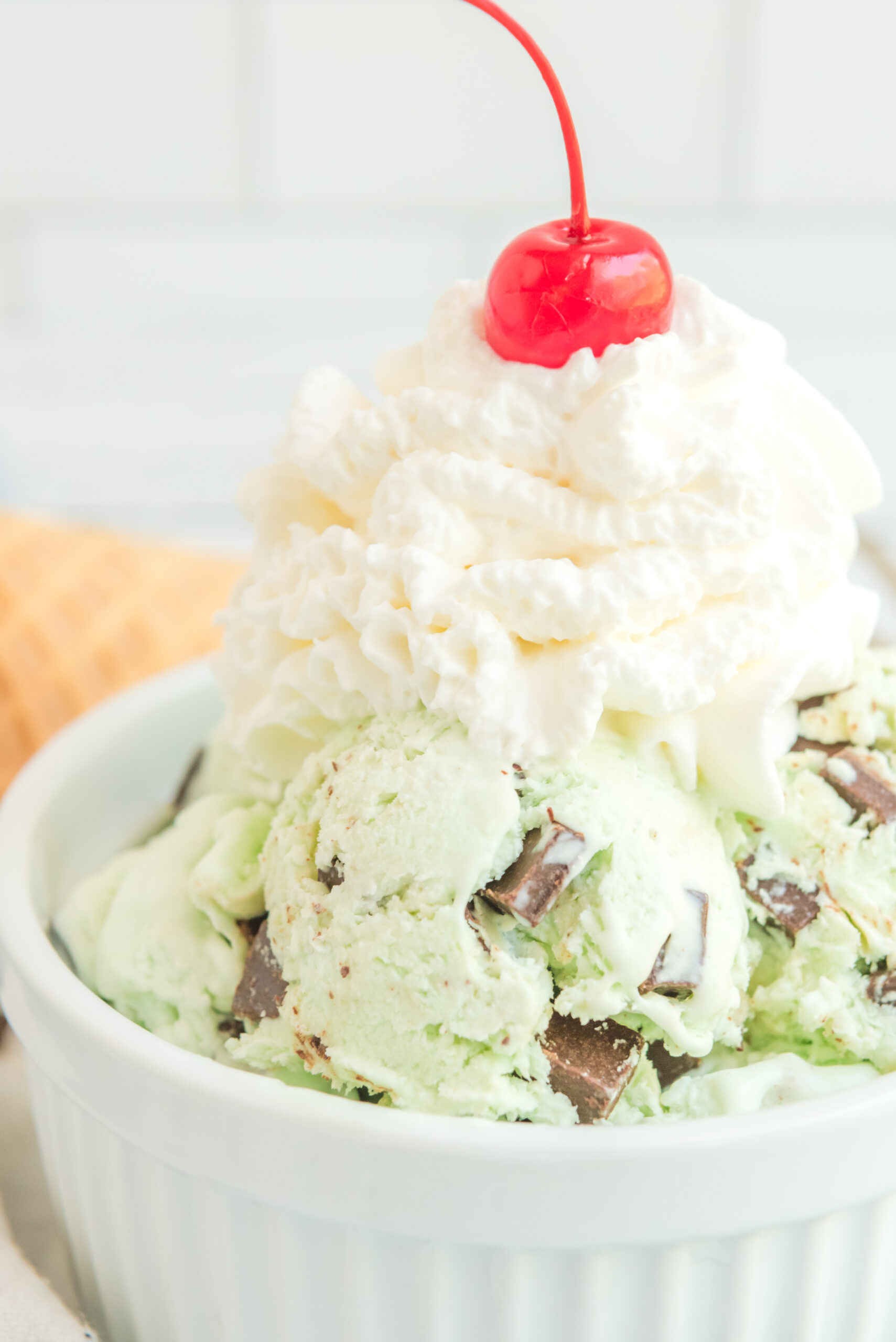 Close up view of scoops of Mint Chocolate Chip Ice Cream in a white bowl garnished with whipped cream and cherry.