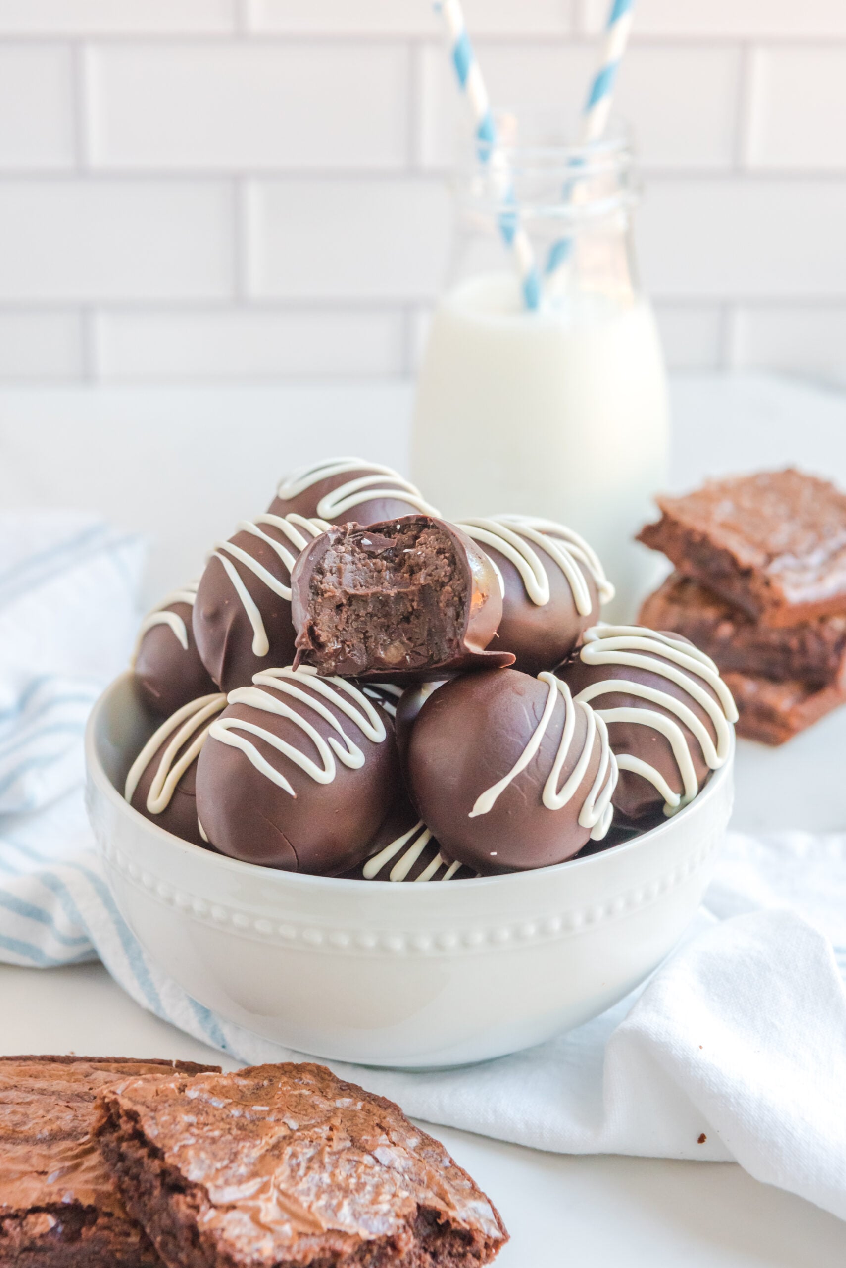 Brownie truffles stacked in a white bowl with brownies and a glass of milk in the background with a blue and white straw.