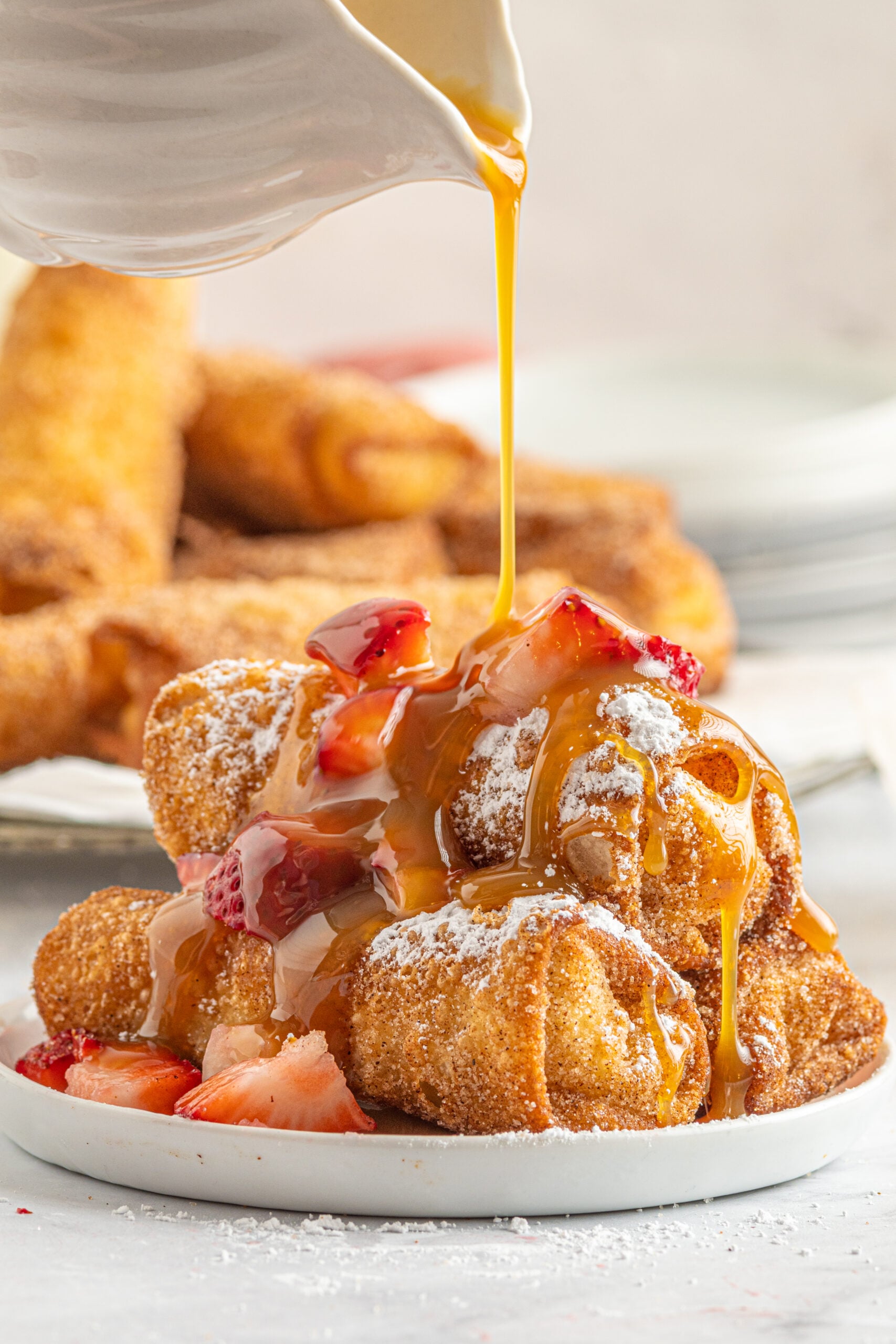 Front view of Cheesecake Egg Rolls stacked on a plate covered with powdered sugar, strawberries and caramel.