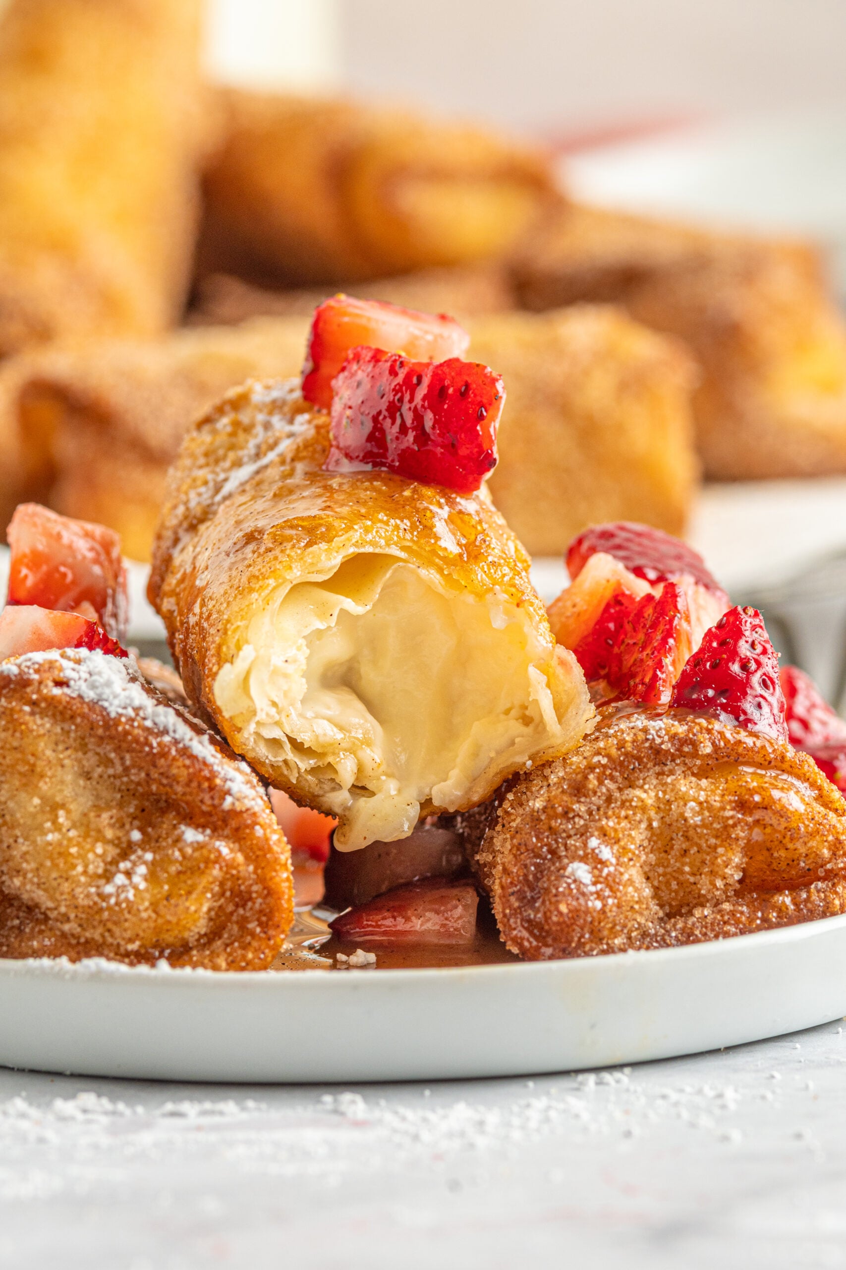 Front view of Cheesecake Egg Rolls stacked on a plate covered with powdered sugar, strawberries and caramel, with a bite removed from the top egg roll.
