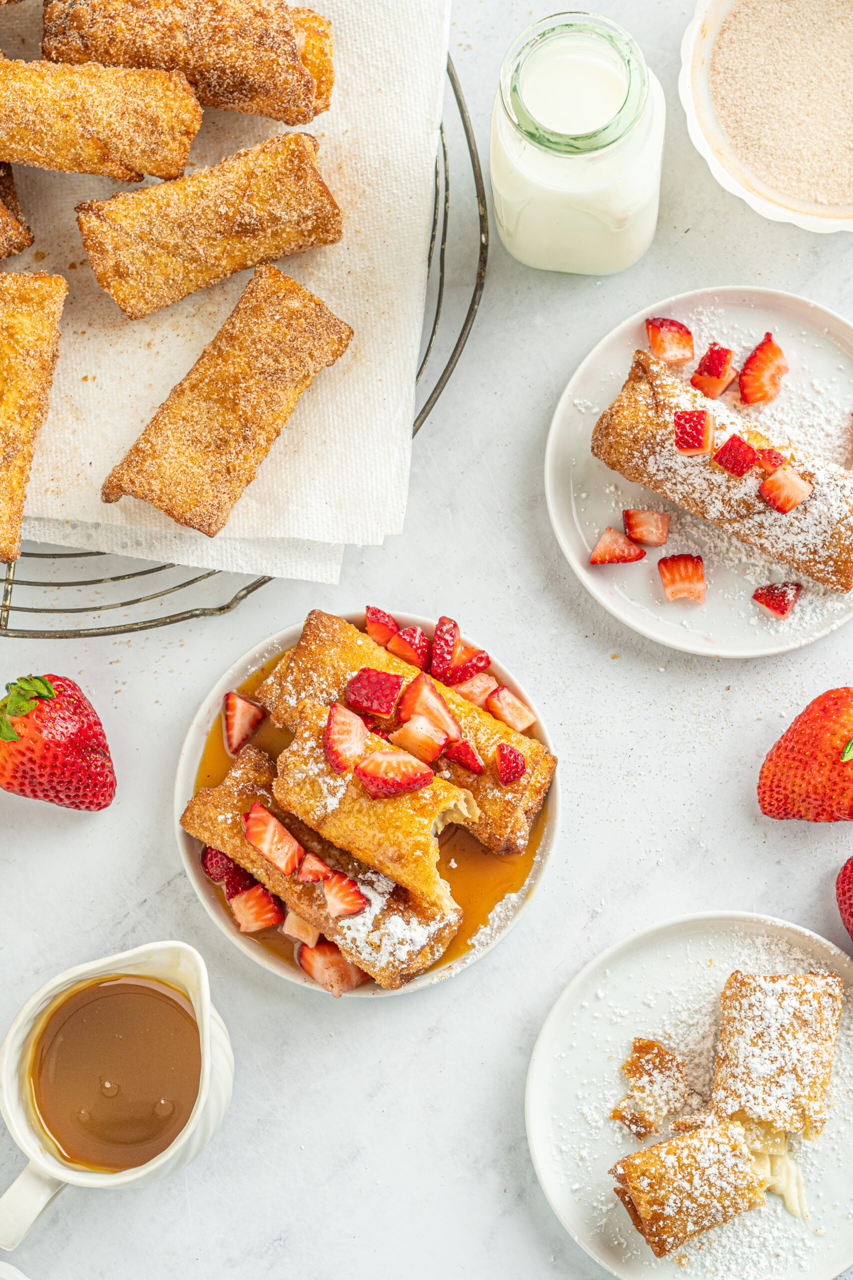 Overhead view of Cheesecake Egg Rolls stacked on a plate covered with powdered sugar, strawberries and caramel, with a bite removed from the top egg roll.