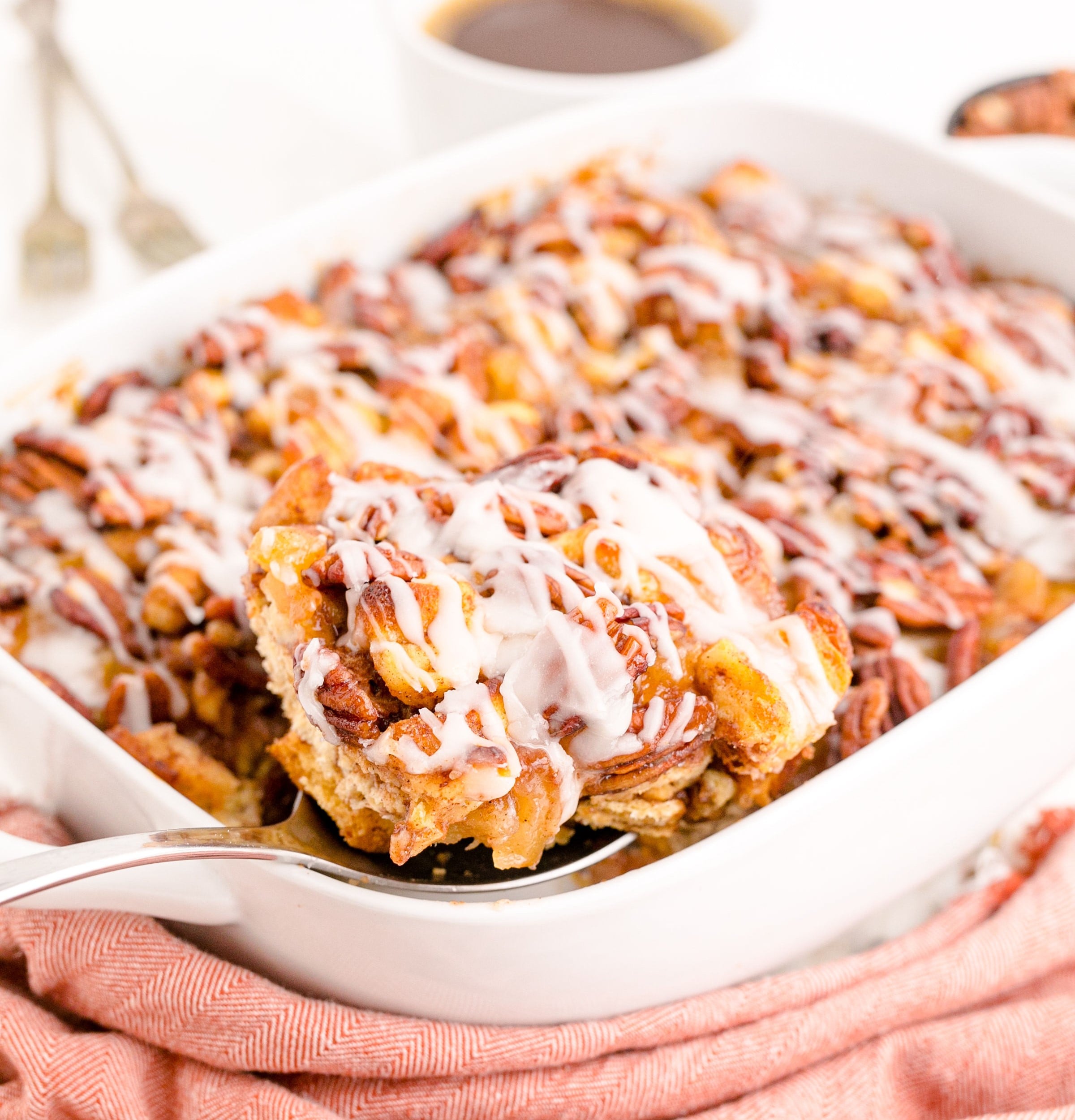 A silver spoon removing a serving of cinnamon roll casserole from a white baking dish.