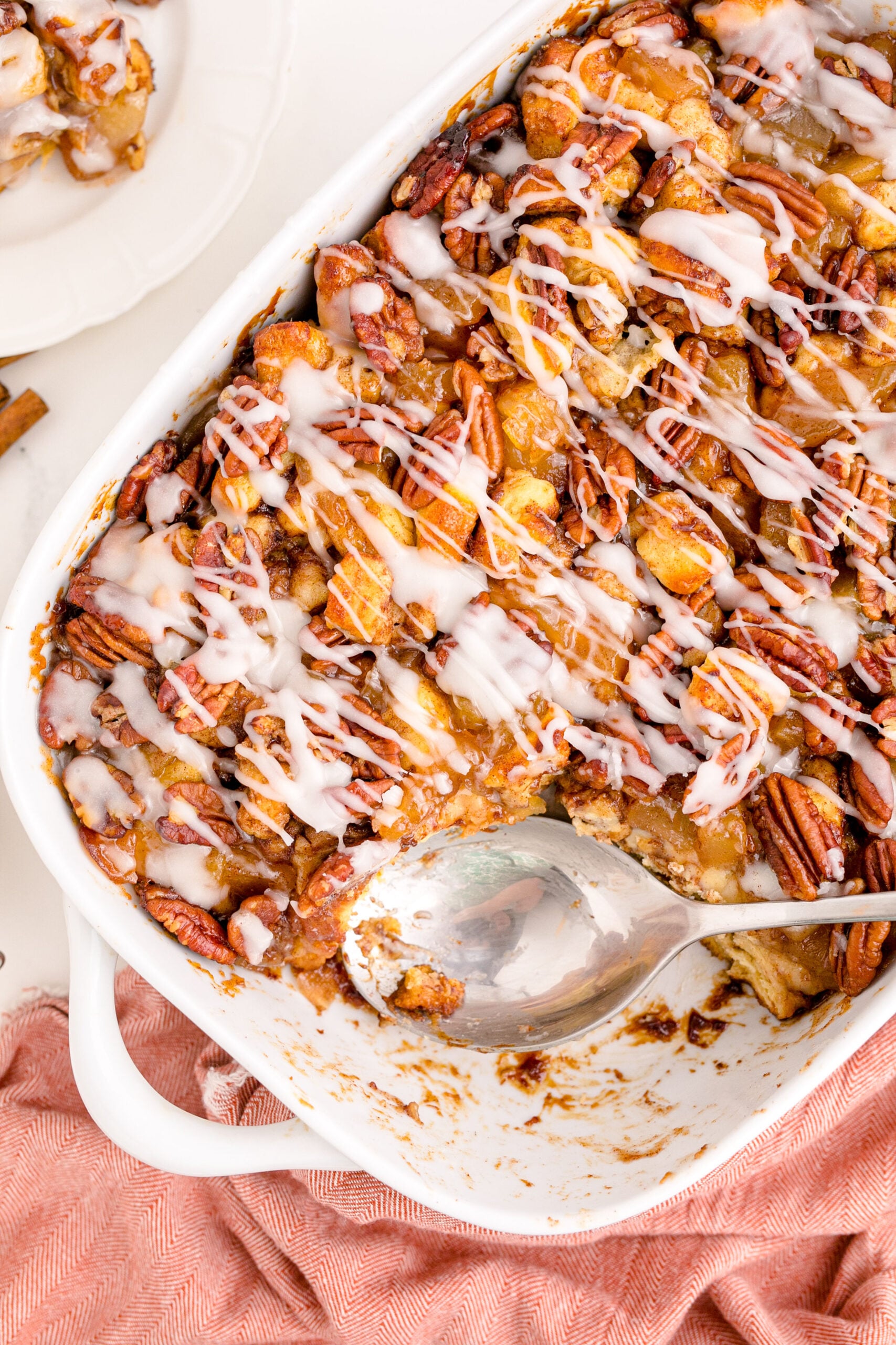Overhead view of A silver spoon removing a serving of cinnamon roll casserole from a white baking dish.