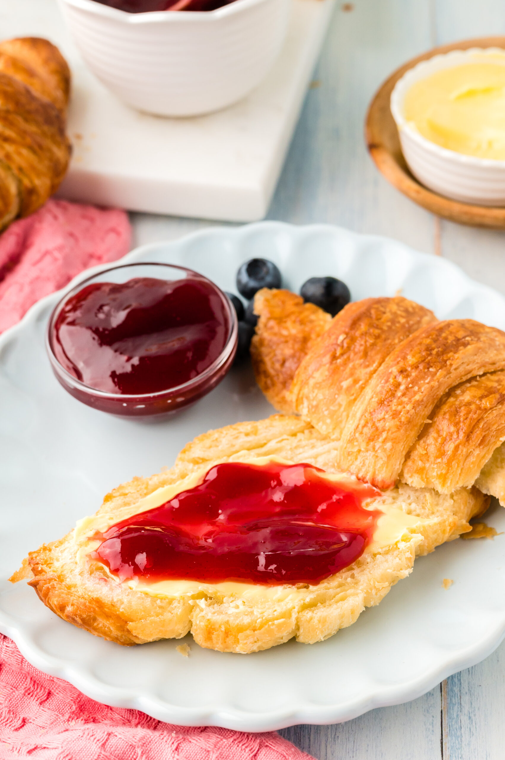 Front view of Homemade Croissants cut in half with jam spread on the bottom half, served on a white dish with jam and fresh blueberries.