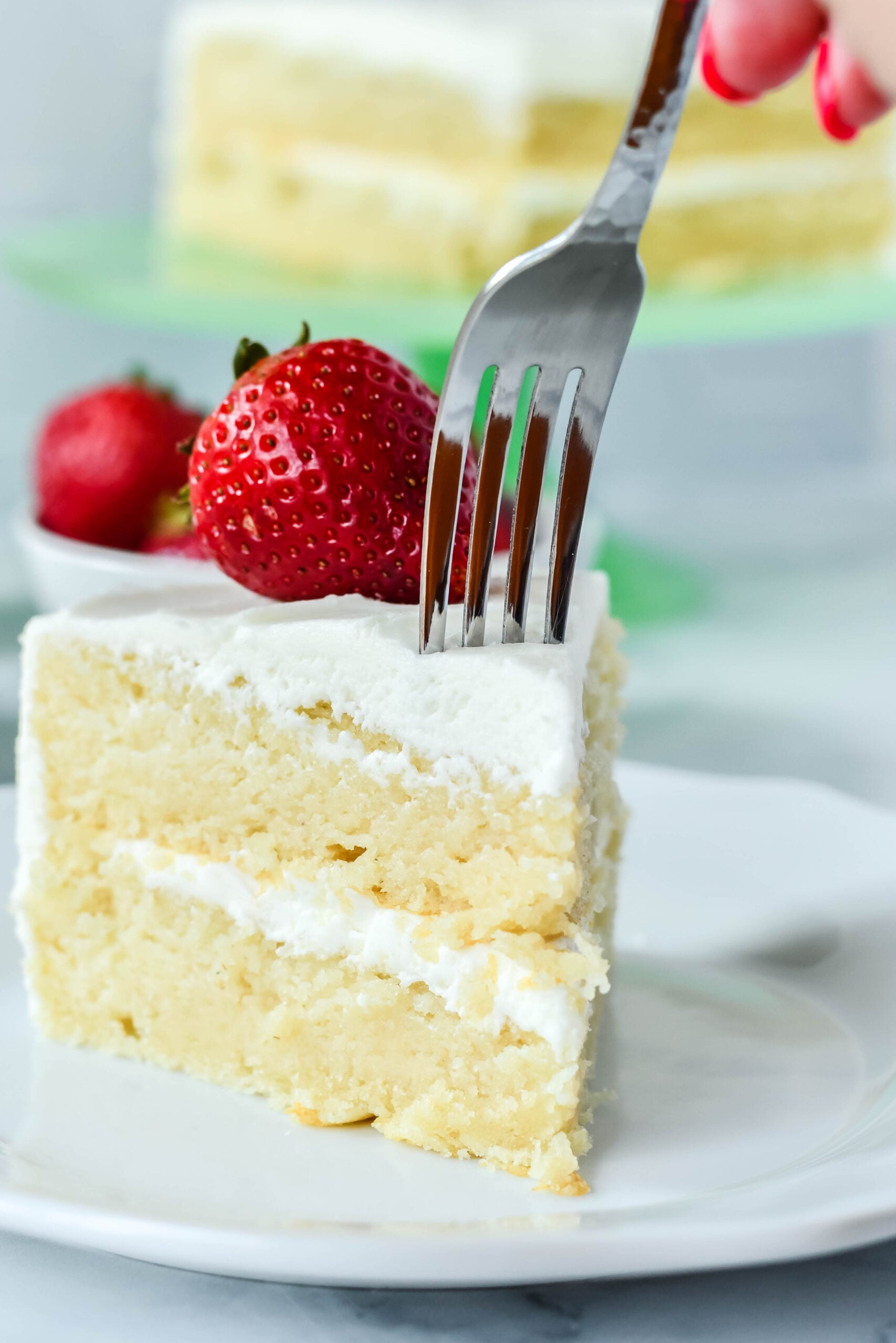Front view of a silver fork removing a bite from a slice of Classic Vanilla Cake on a white plate garnished with a strawberry.