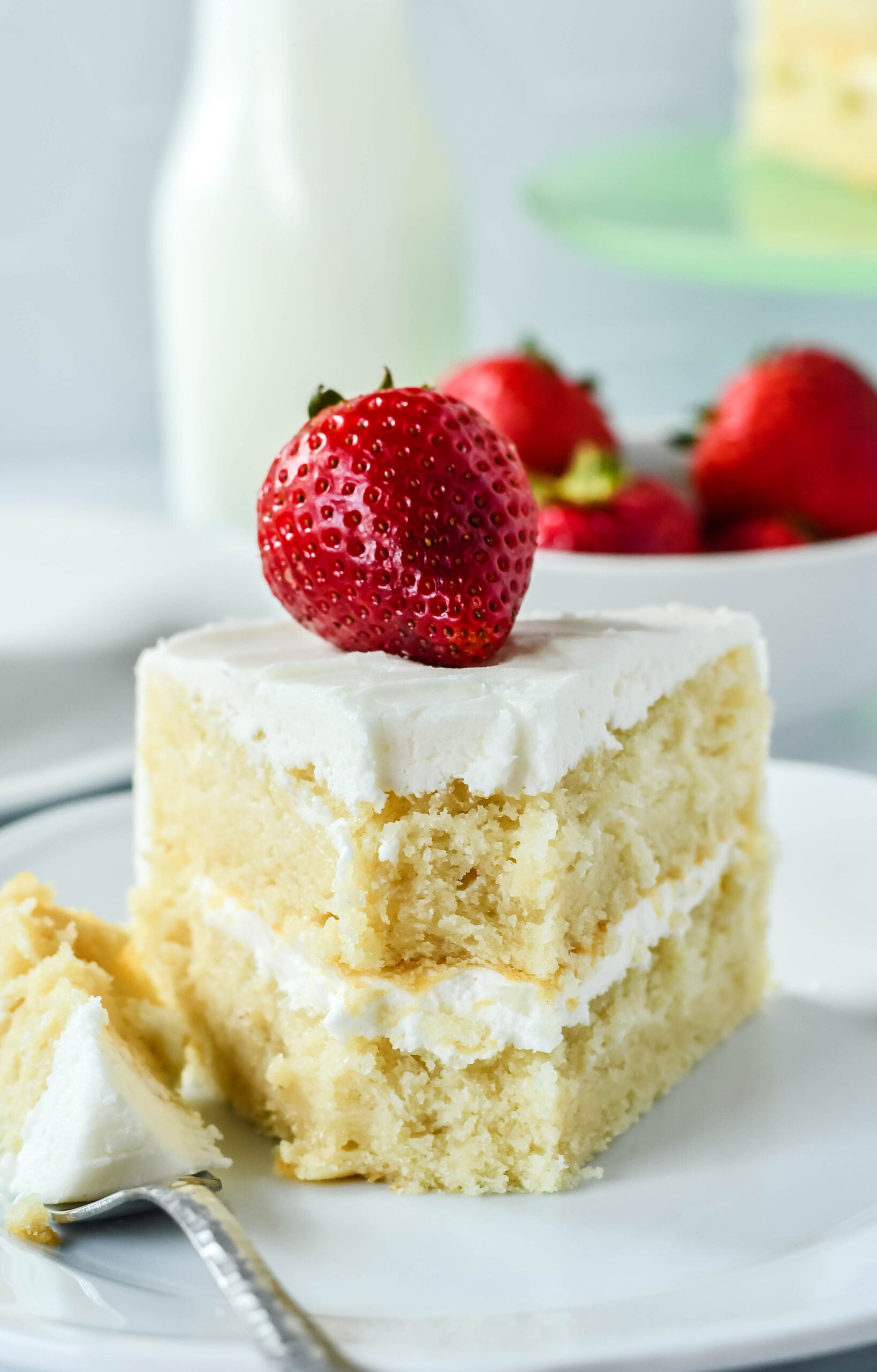Up close view of a slice of Classic Vanilla Cake on a white plate garnished with a strawberry with a bite removed.