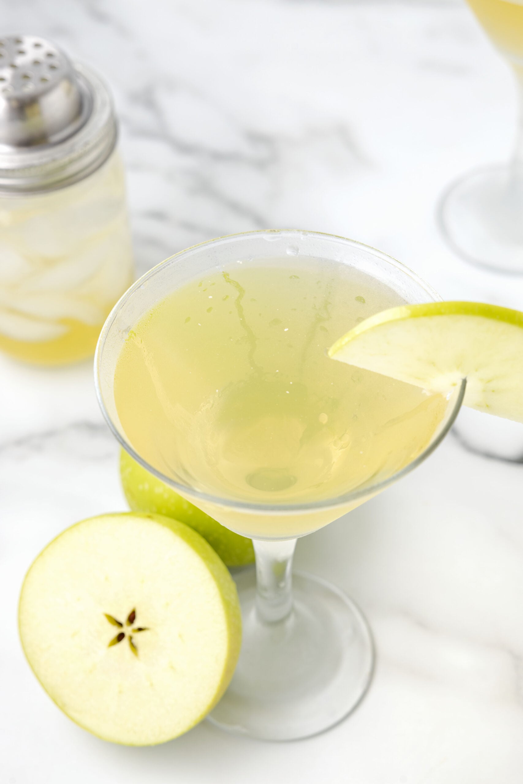 An Appletini in a martini glass garnished with a sliced apple with apples in the background.