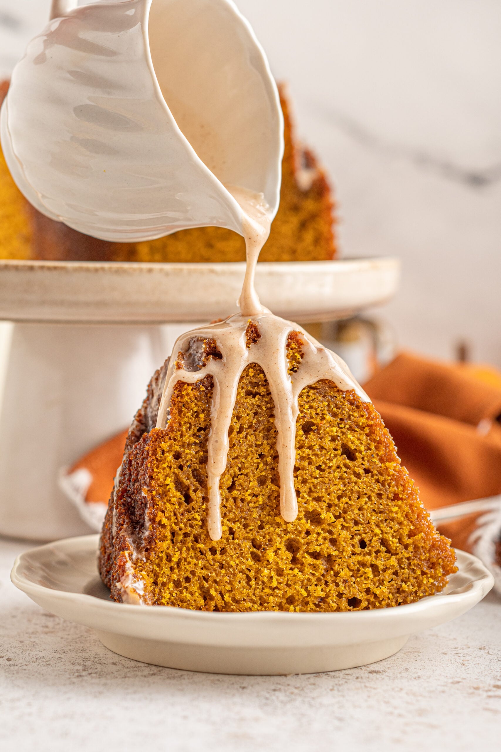 A slice of Pumpkin Bundt Cake with Cinnamon Sugar Glaze on a white dish with glaze being poured over top.