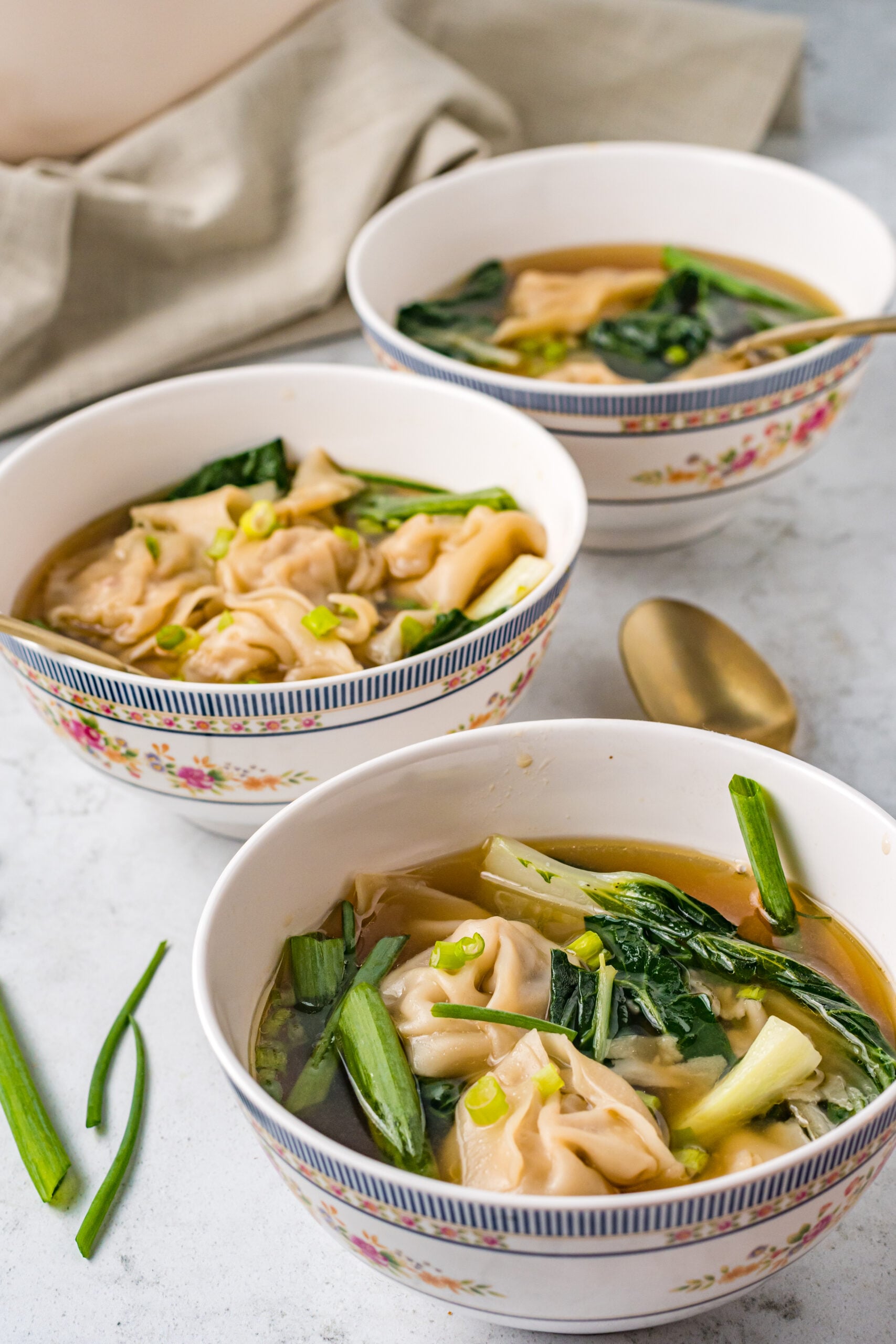 Side view of Wonton Soup in three white bowls topped with sliced green onion.