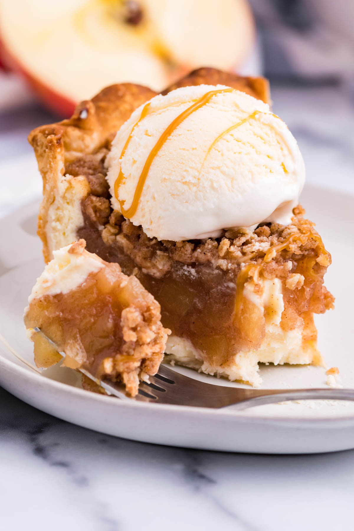 Front view of a slice of Apple Crumb Cheesecake Pie on a white dish with a scoop of vanilla ice cream with a bite removed by a silver fork.