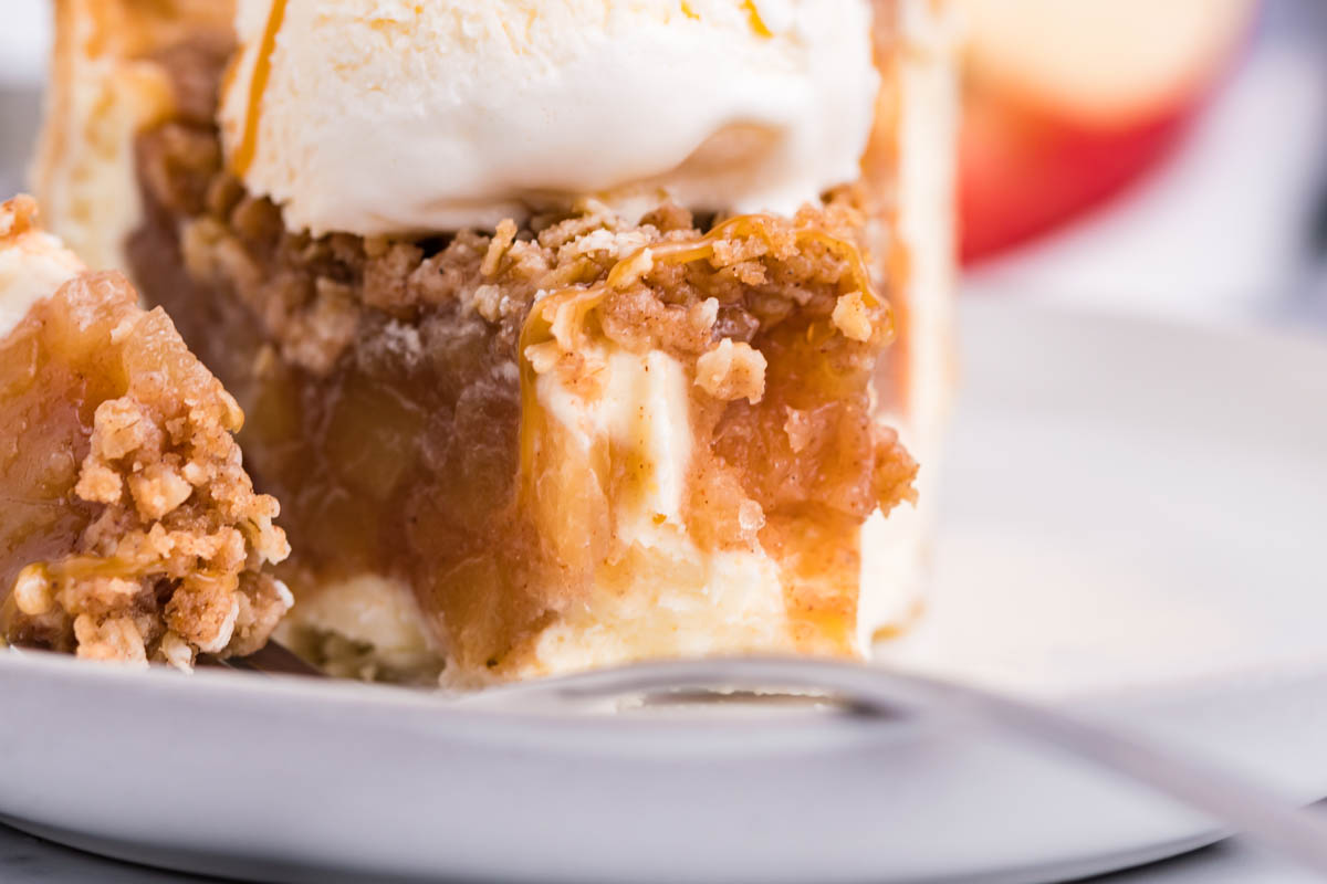 Front view of a slice of Apple Crumb Cheesecake Pie on a white dish with a scoop of vanilla ice cream with a bite removed by a silver fork.
