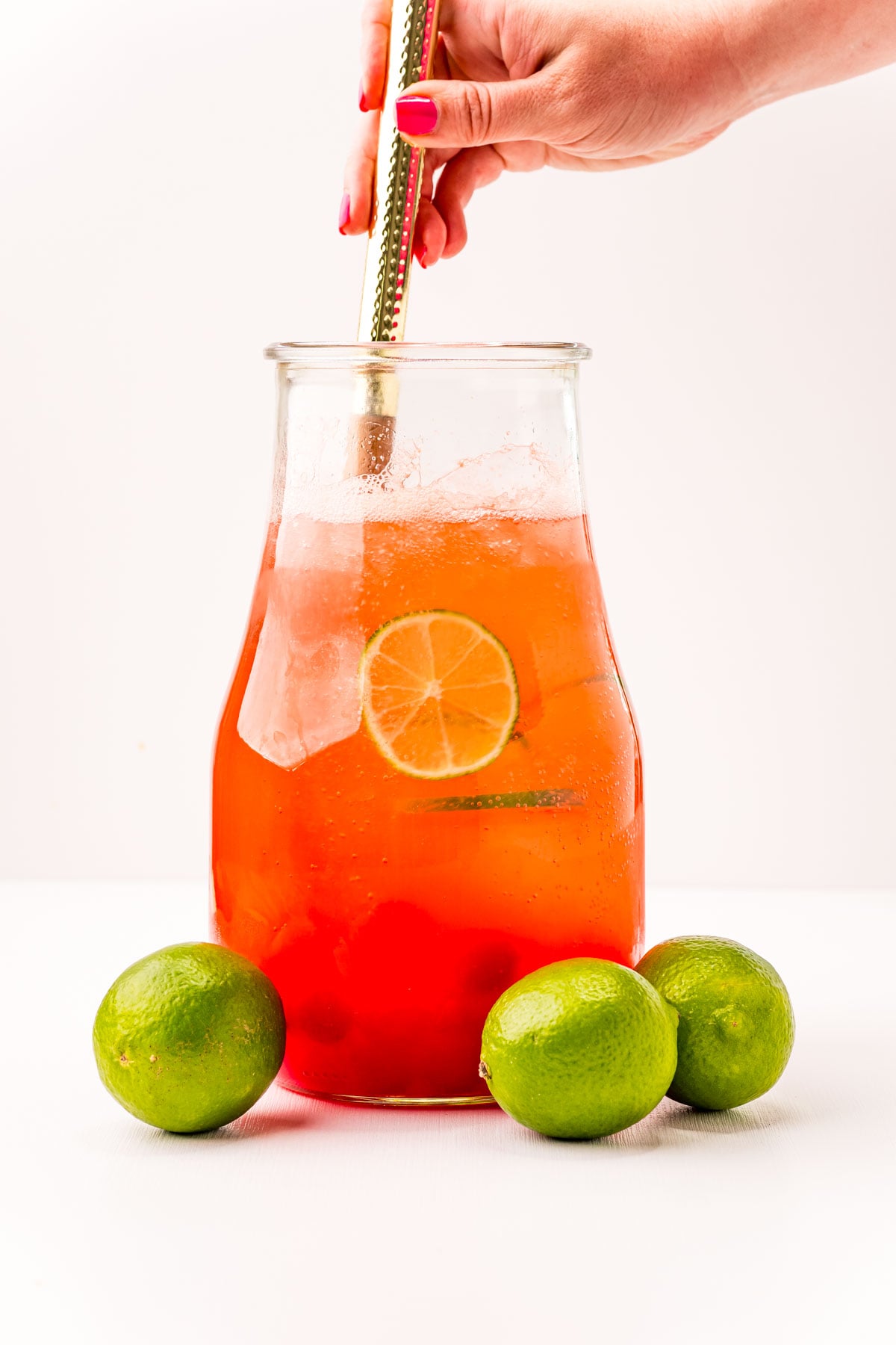 Front view of Cherry Limeade in a glass pitcher surrounded by whole limes.
