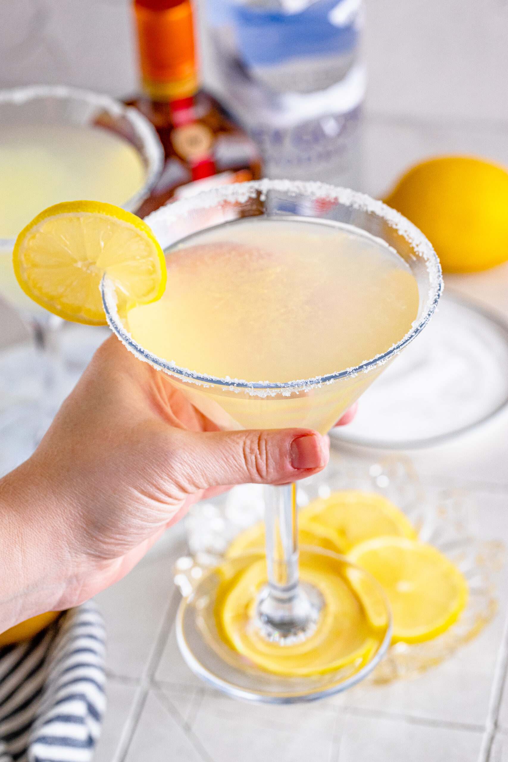 A hand holding a lemon drop martini with lemon slices in the background. 