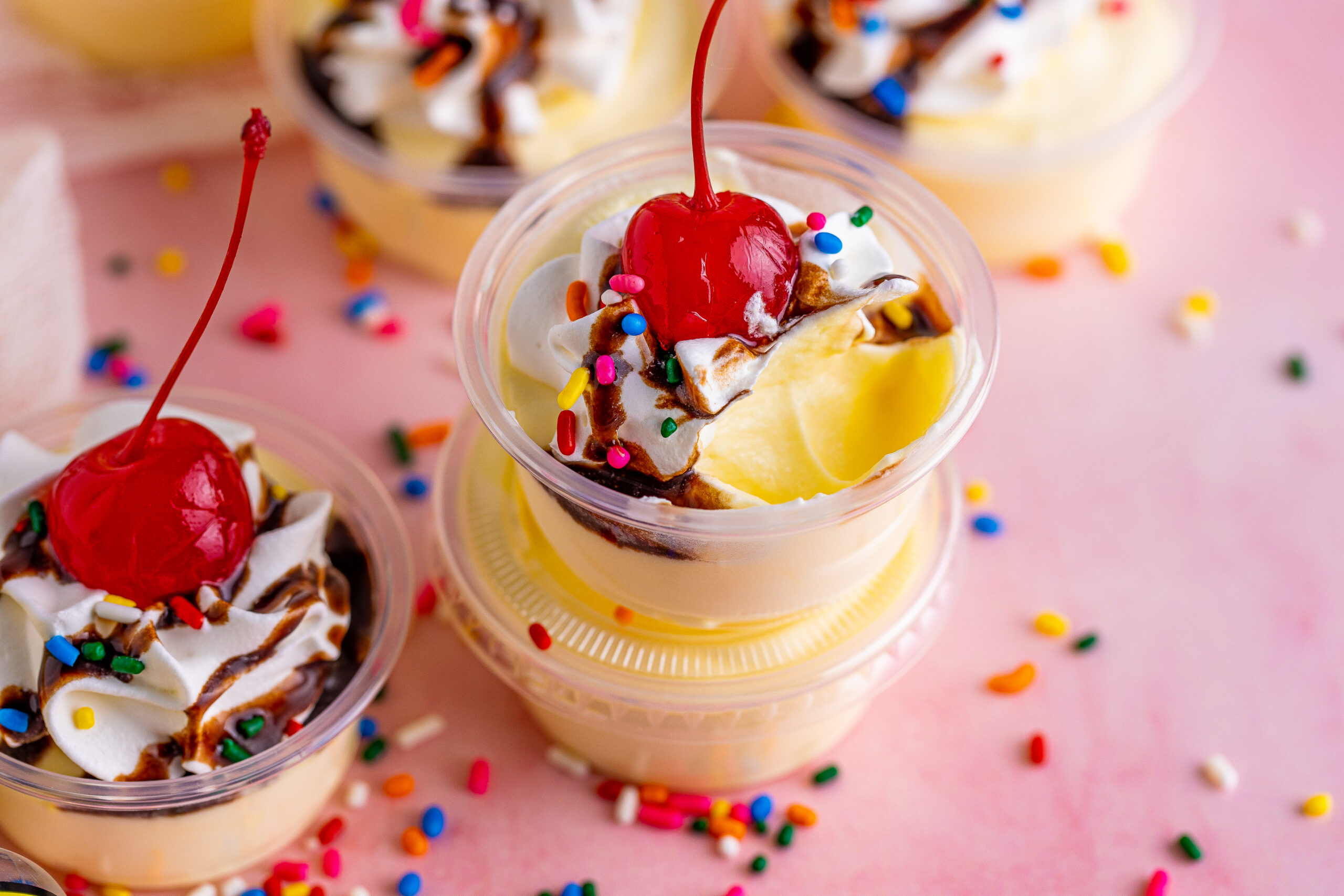 Banana pudding shots with a scoop missing topped with a cherry, whipped cream and chocolate sauce