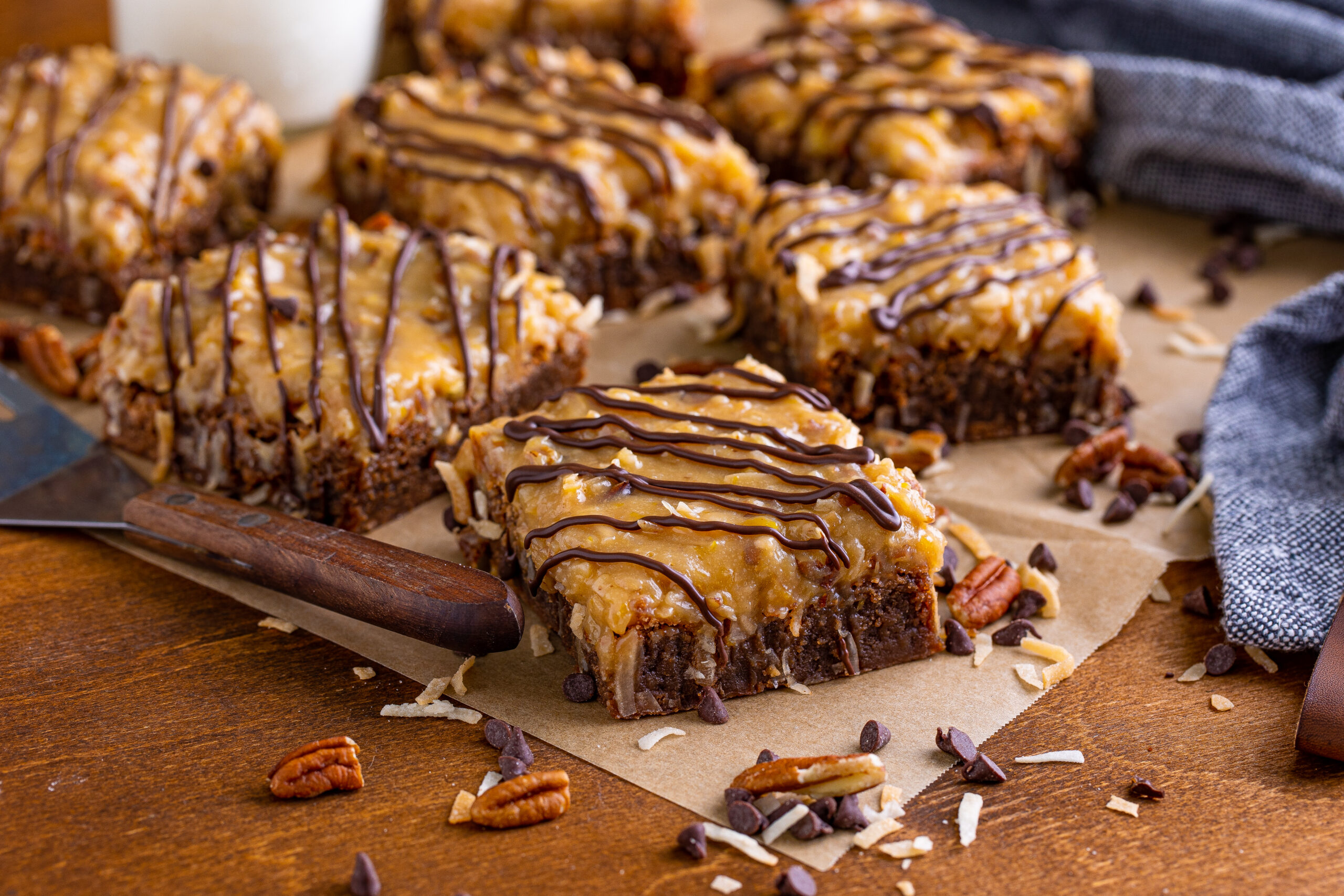 German Chocolate Brownies on a wood table surrounded by chopped pecans and chocolate chips 