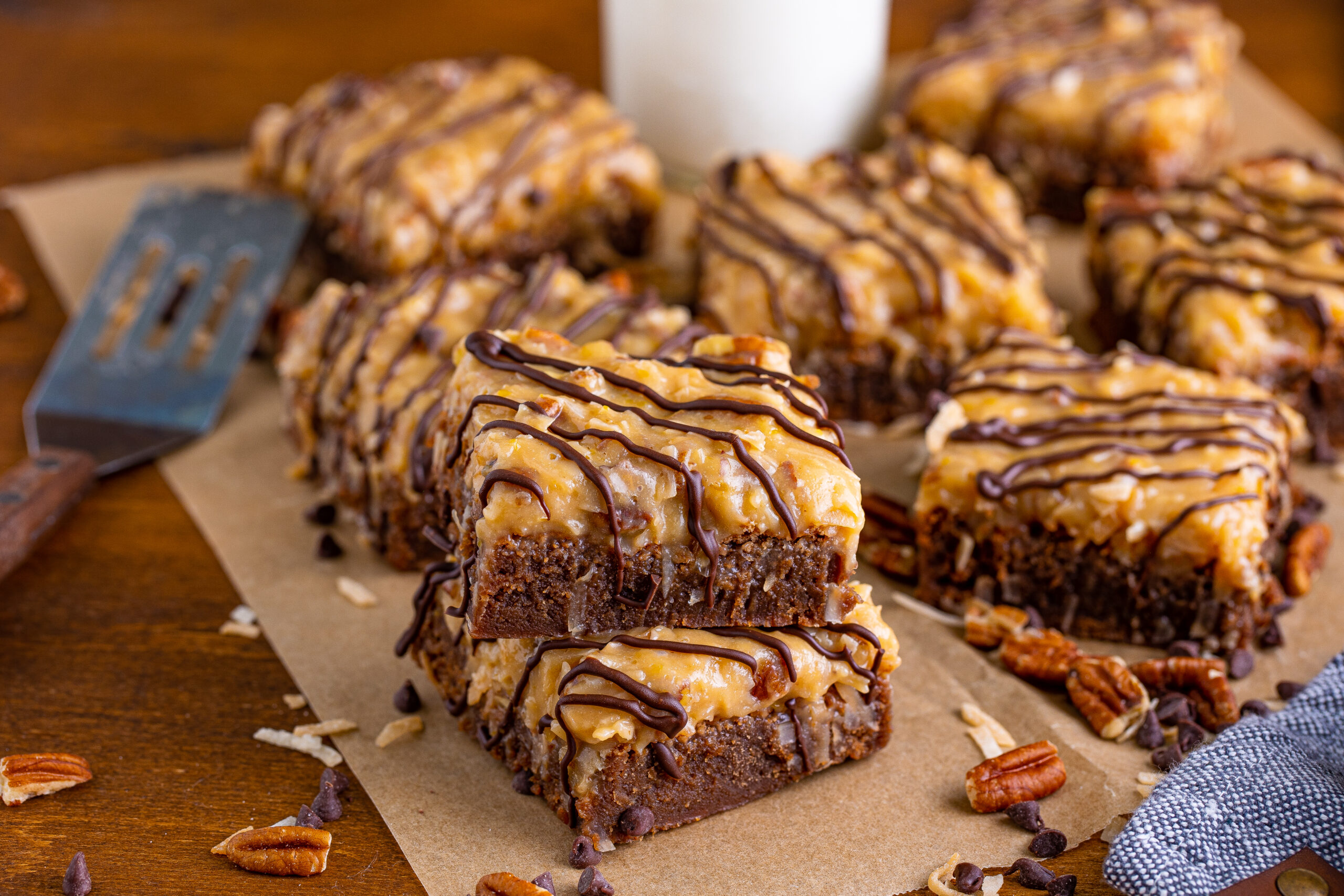 German Chocolate Brownies on a wood table surrounded by chopped pecans and chocolate chips 