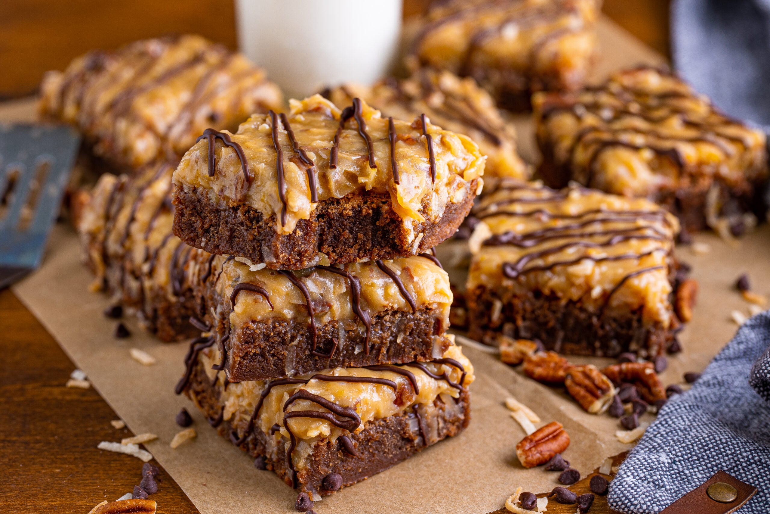 Three stacked German Chocolate Brownies with a bite missing, on a wood table surrounded by chopped pecans and chocolate chips 