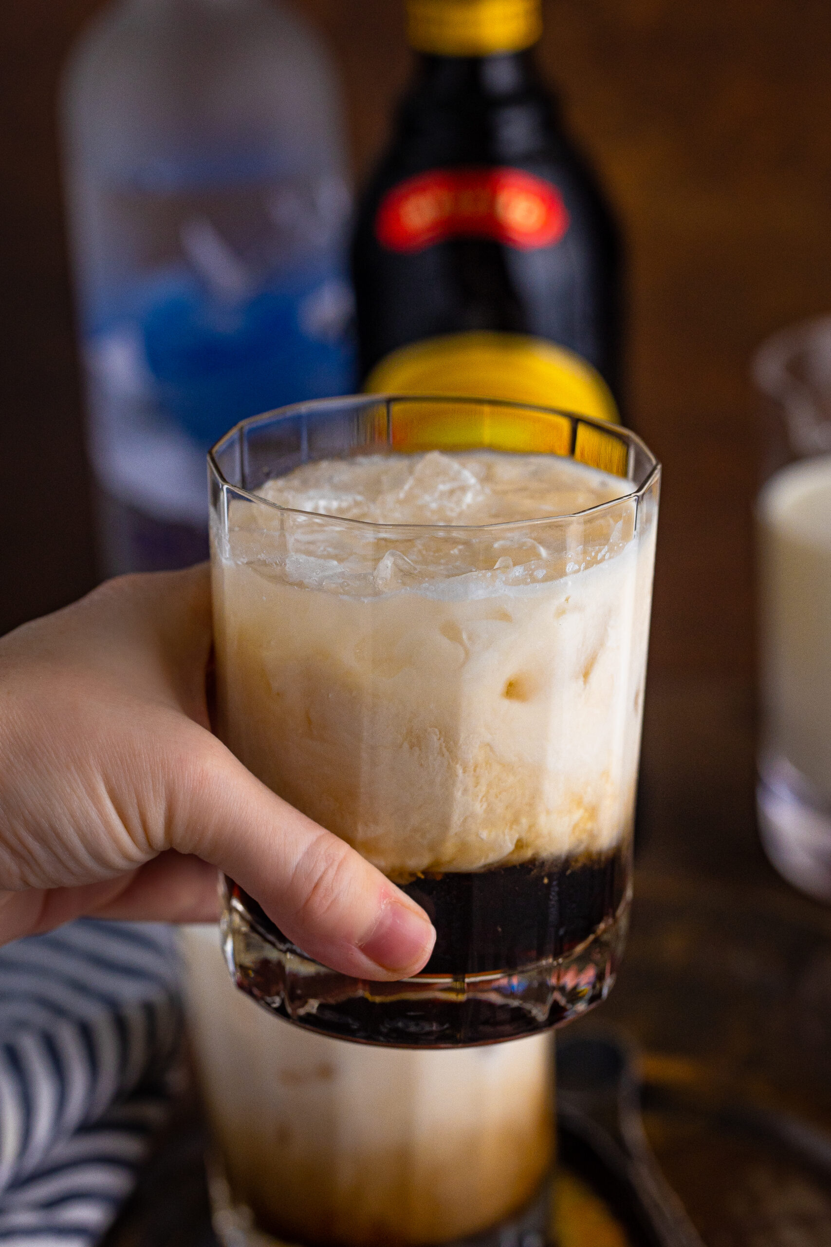 A hand holding a White Russian drink in a clear glass.