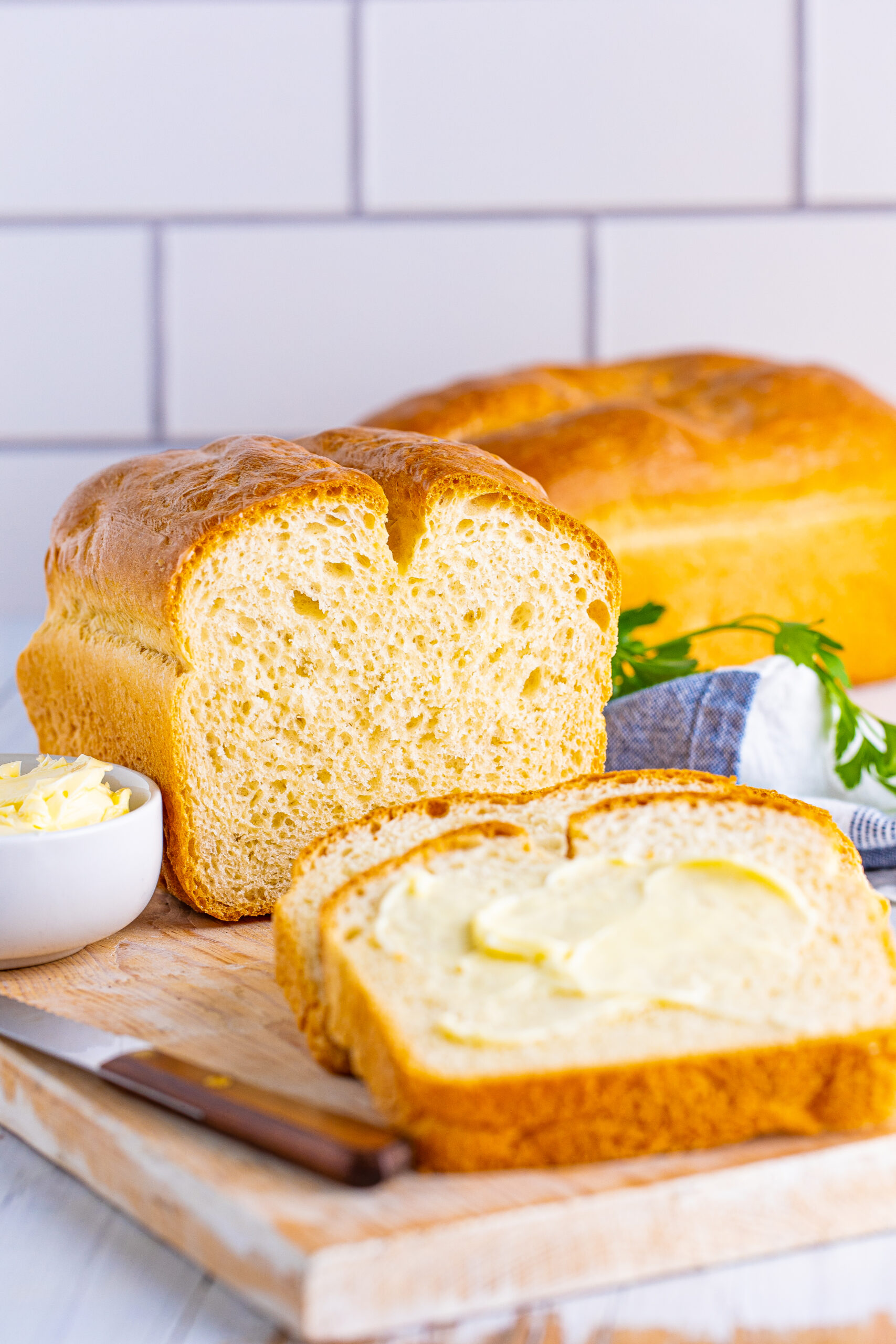 Front view of a loaf of White Bread with two slices removed and buttered on a wooden cutting board.
