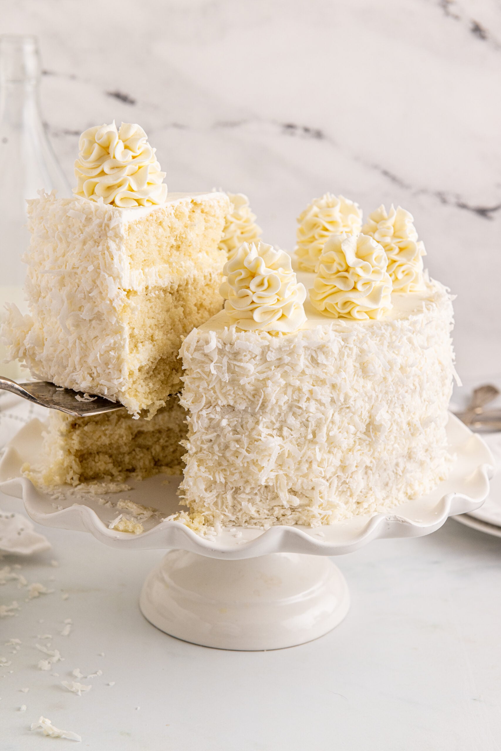 Front view of Coconut Layer Cake on a white cake stand with a slice being removed by a silver cake server.
