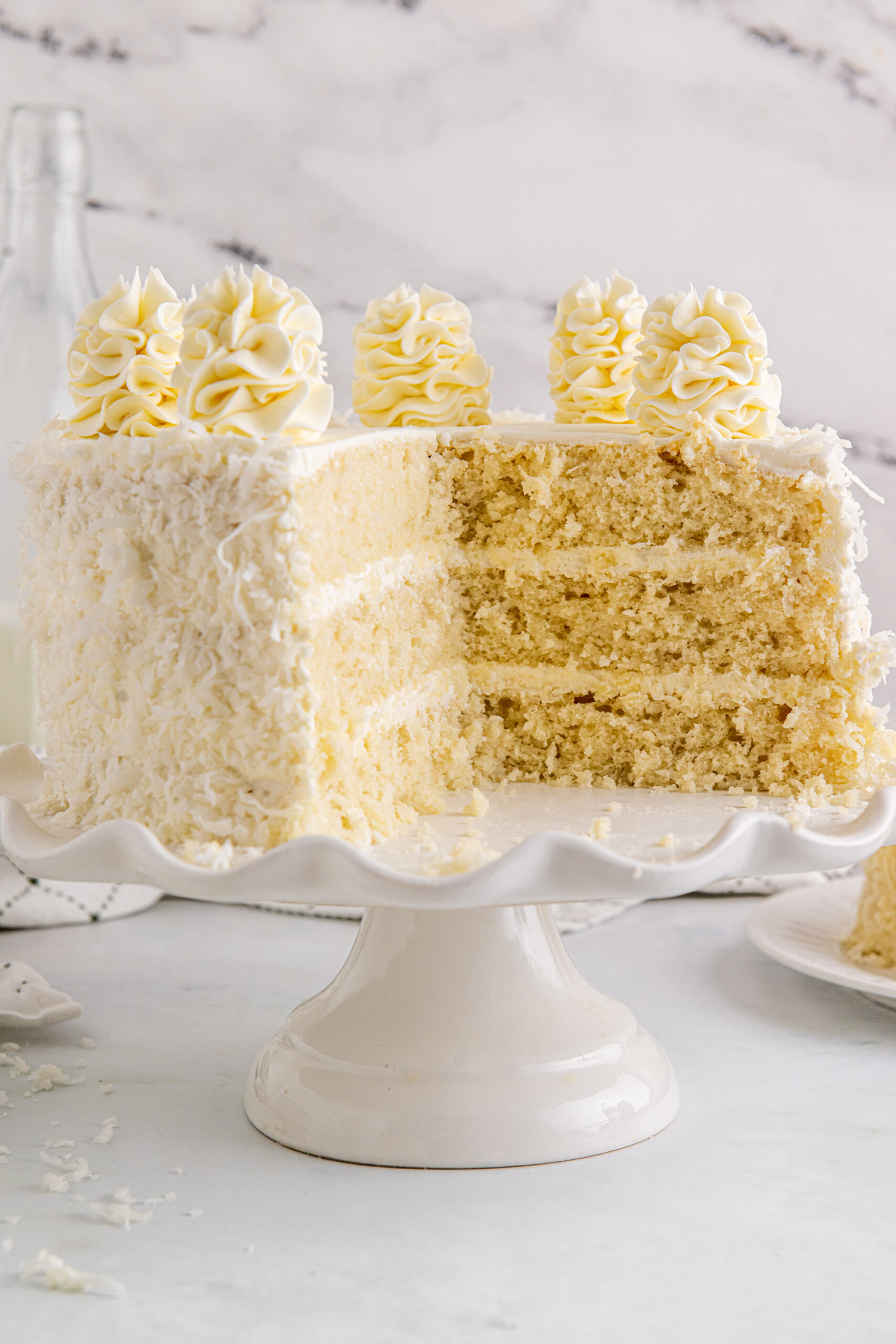 Front view of Coconut Layer Cake on a white cake stand with two slices removed.