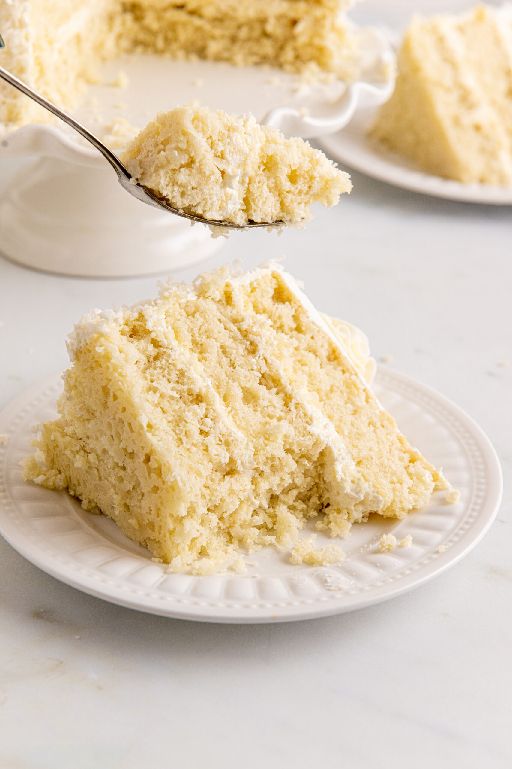 Front view of a slice of Coconut Layer Cake on a white plate with a bite removed by a silver fork.
