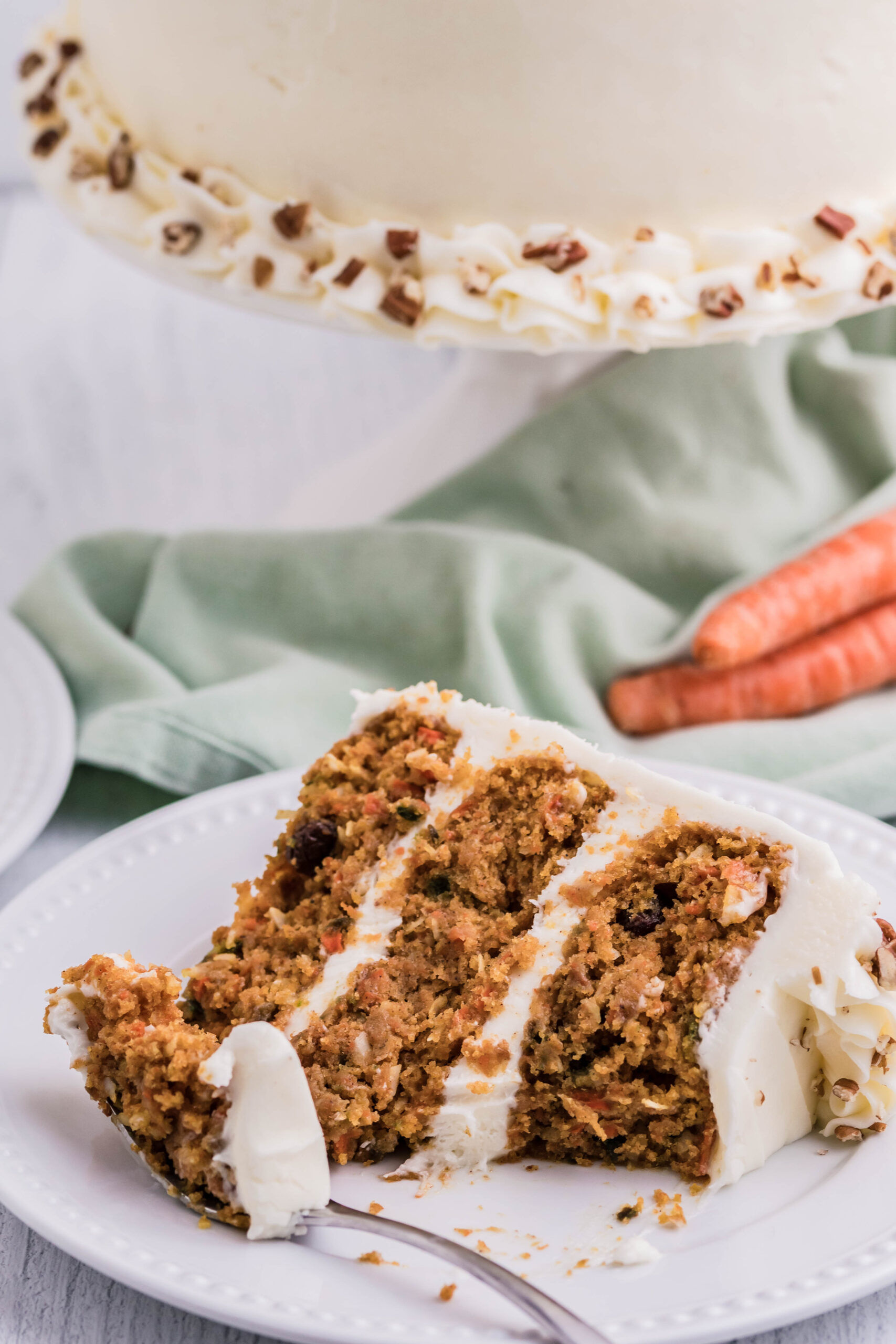 Front view of a slice of Carrot Cake served on a white plate with a bite removed by a silver fork.