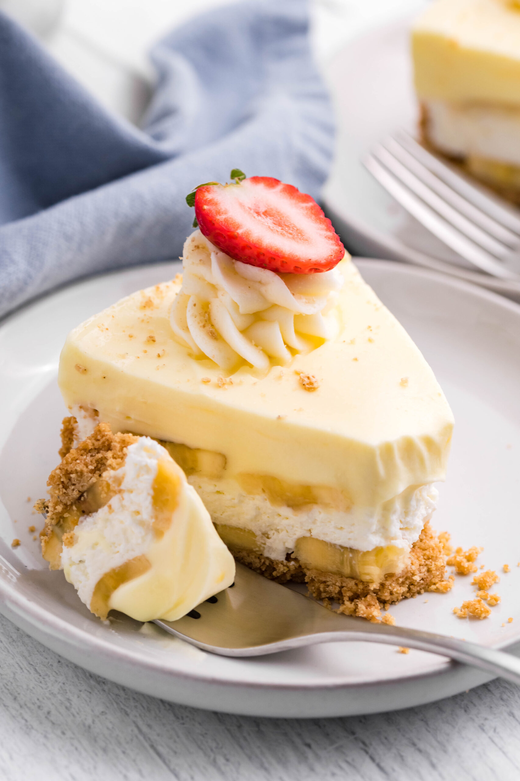 Side view of a slice of Banana Cream Cheesecake on a white plate with a bite removed by a silver fork.