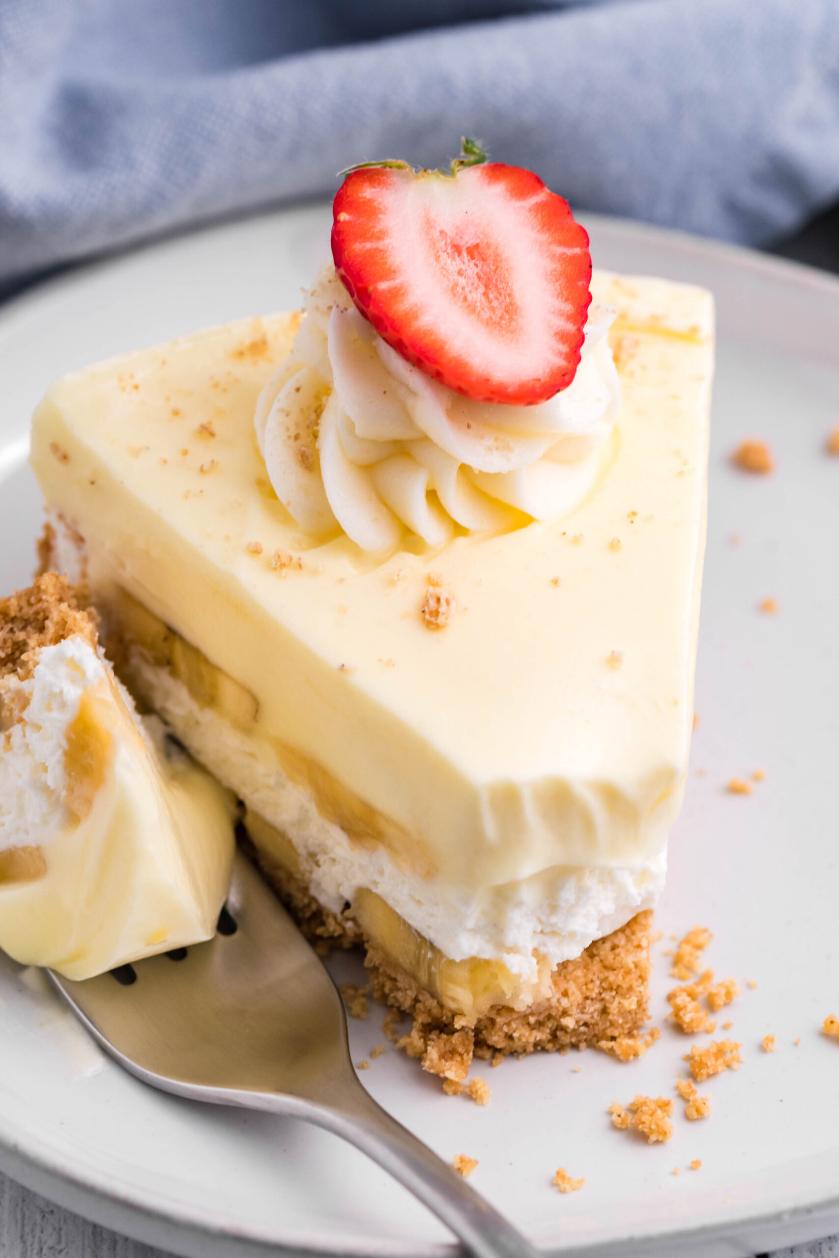 Front view of a slice of Banana Cream Cheesecake on a white plate with a bite removed by a silver fork.
