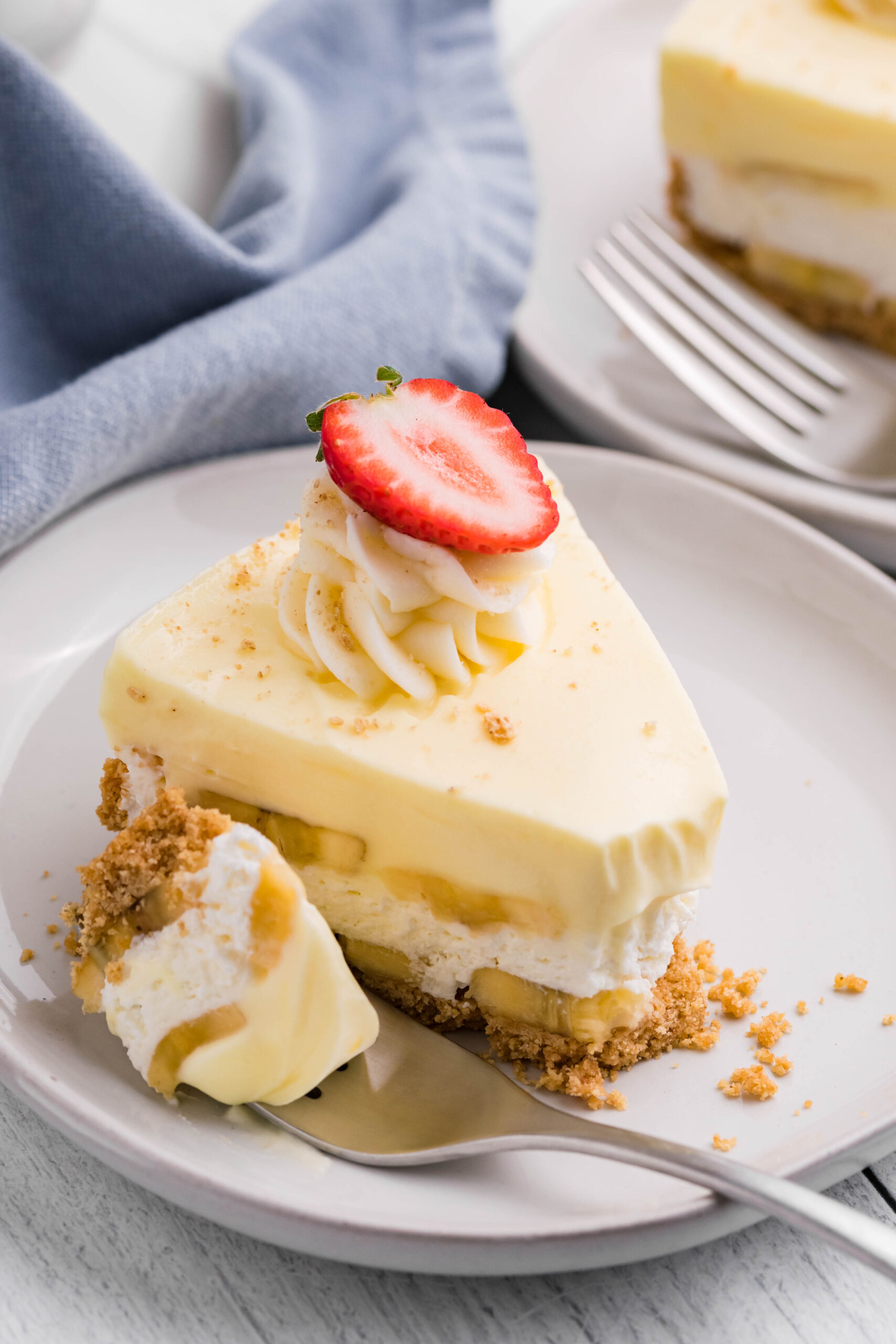 Side view of a slice of Banana Cream Cheesecake on a white plate with a bite removed by a silver fork.