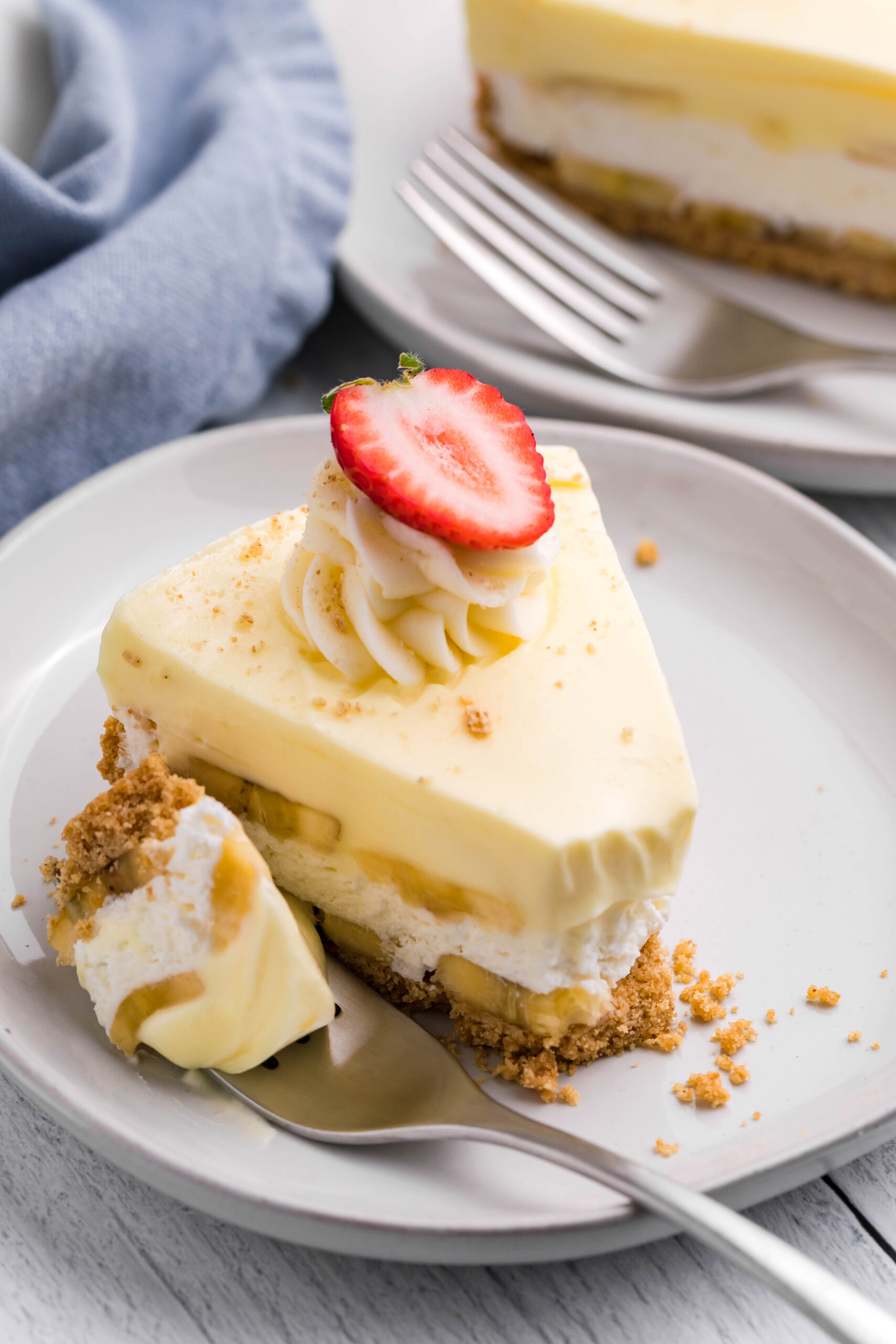 Front view of a slice of Banana Cream Cheesecake on a white plate with a bite removed by a silver fork.