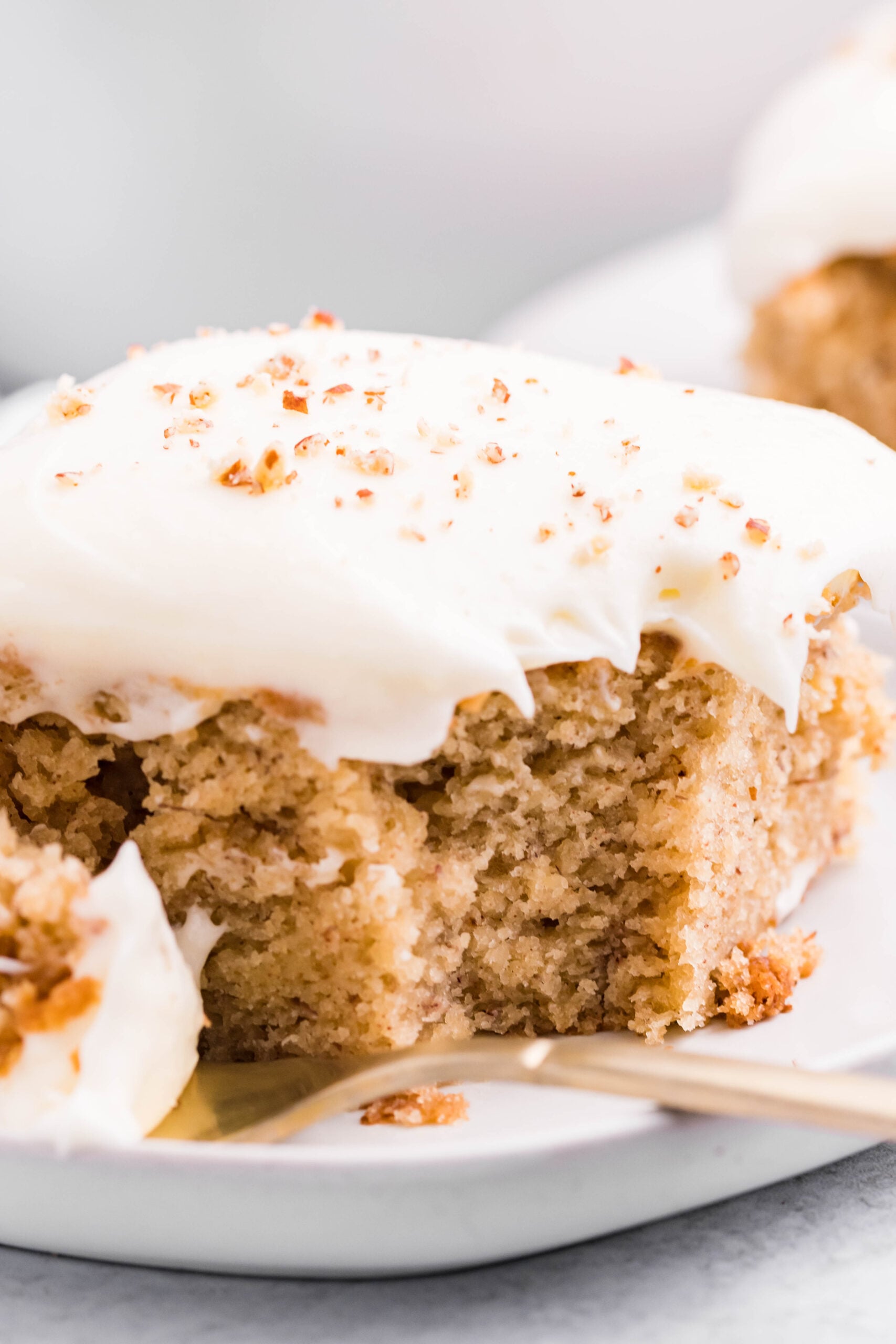 Close up view of a slice of Banana Sheet Cake on a white plate with a bite removed by a gold fork.