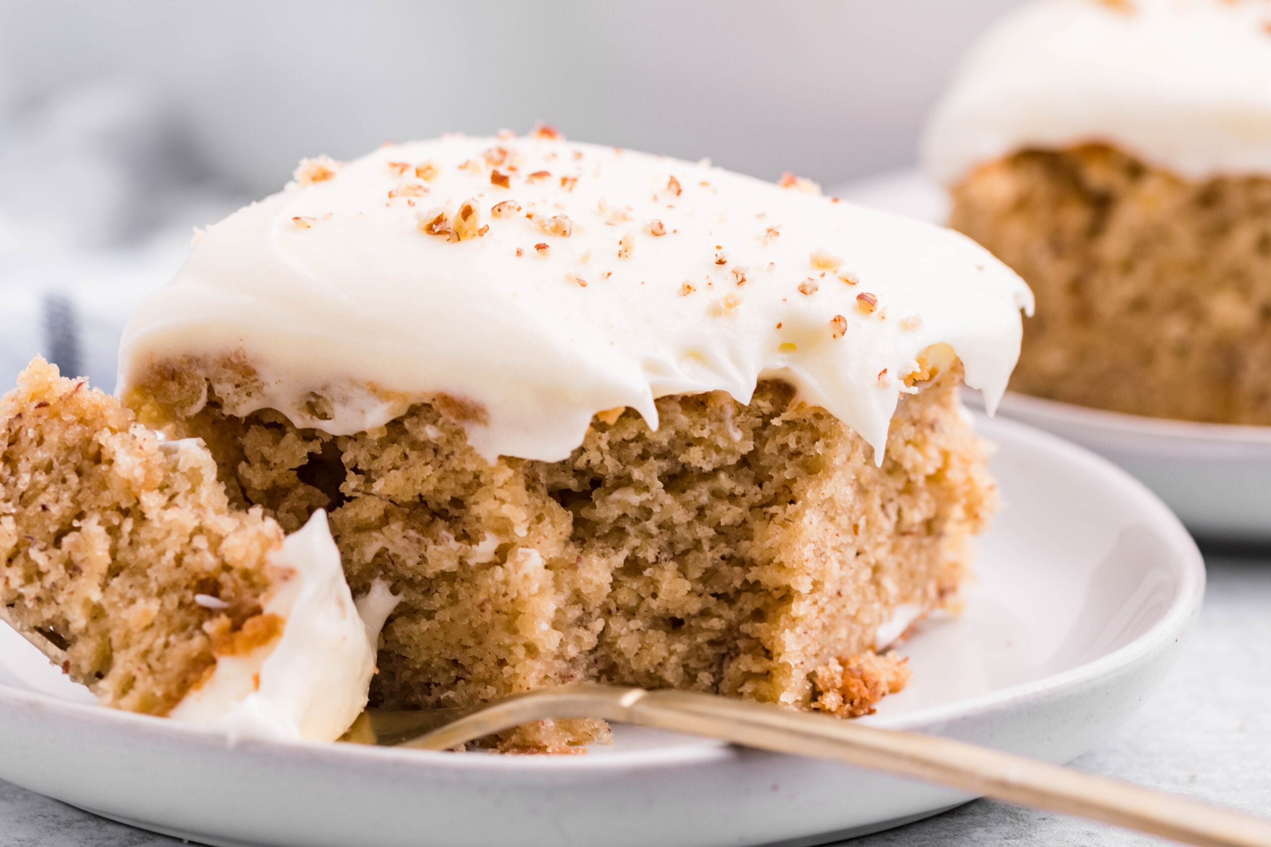 Front view of a slice of Banana Sheet Cake on a white plate with a bite removed by a gold fork.