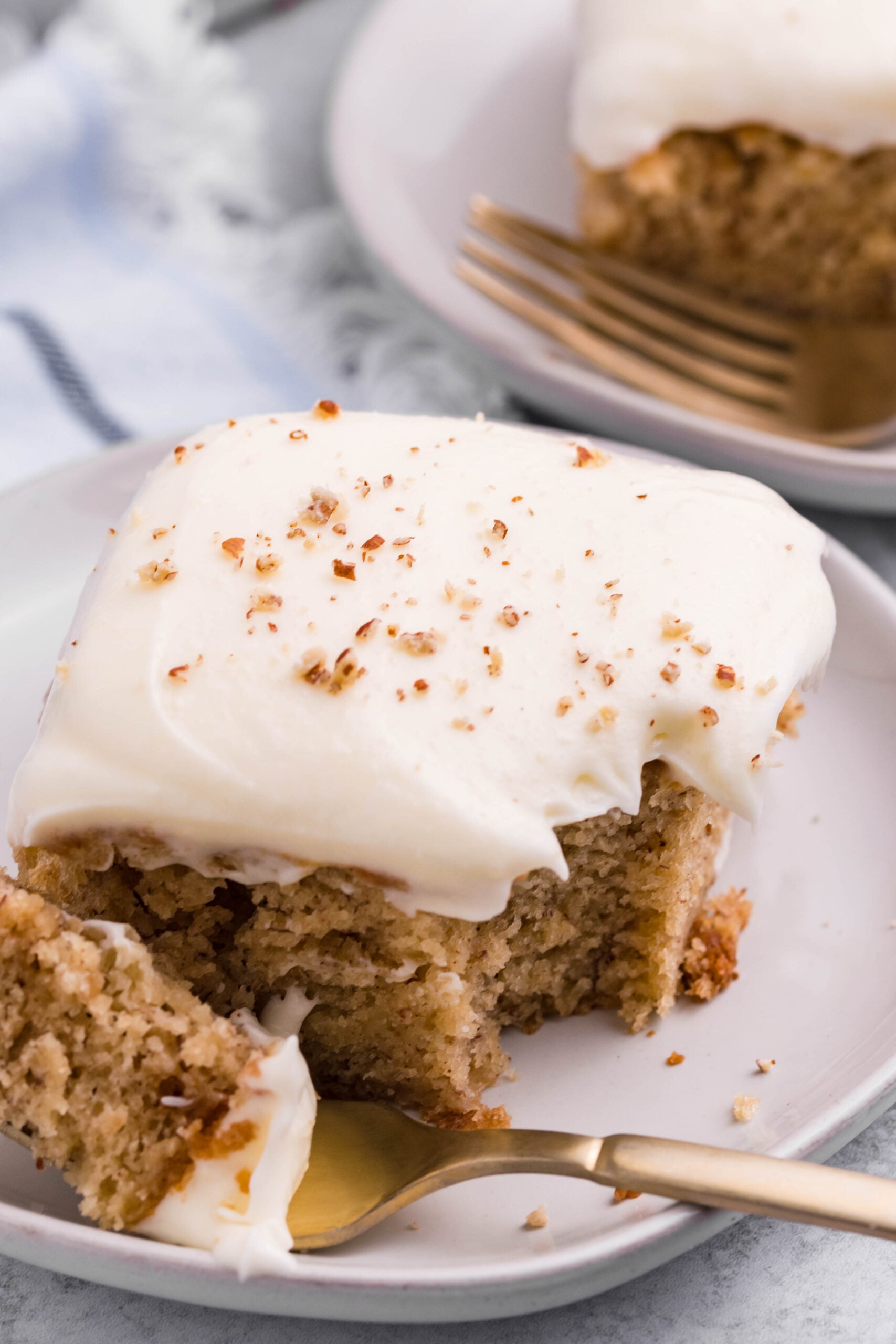 Close up view of a slice of Banana Sheet Cake on a white plate with a bite removed by a gold fork.