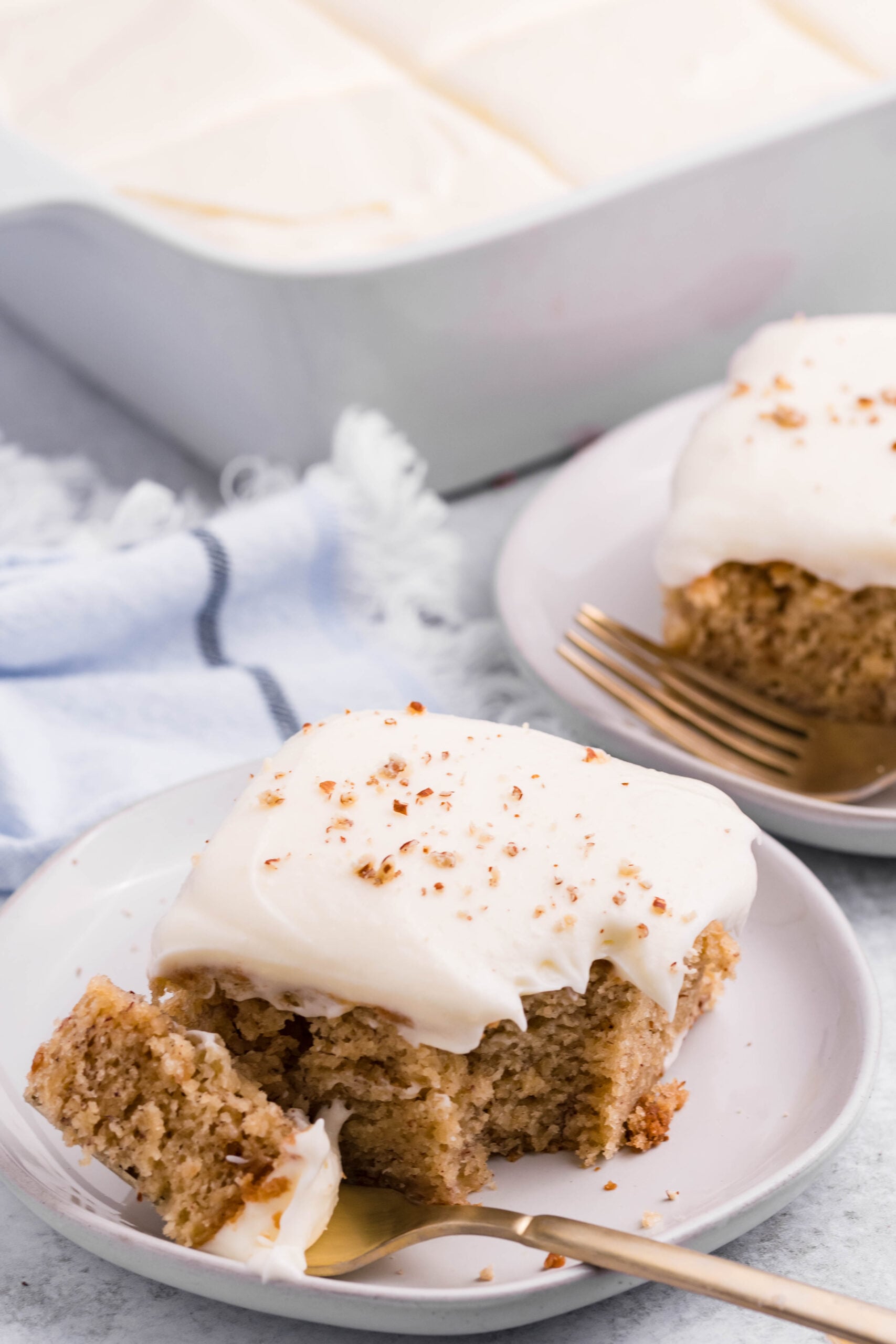 Front view of a slice of Banana Sheet Cake on a white plate with a bite removed by a gold fork.