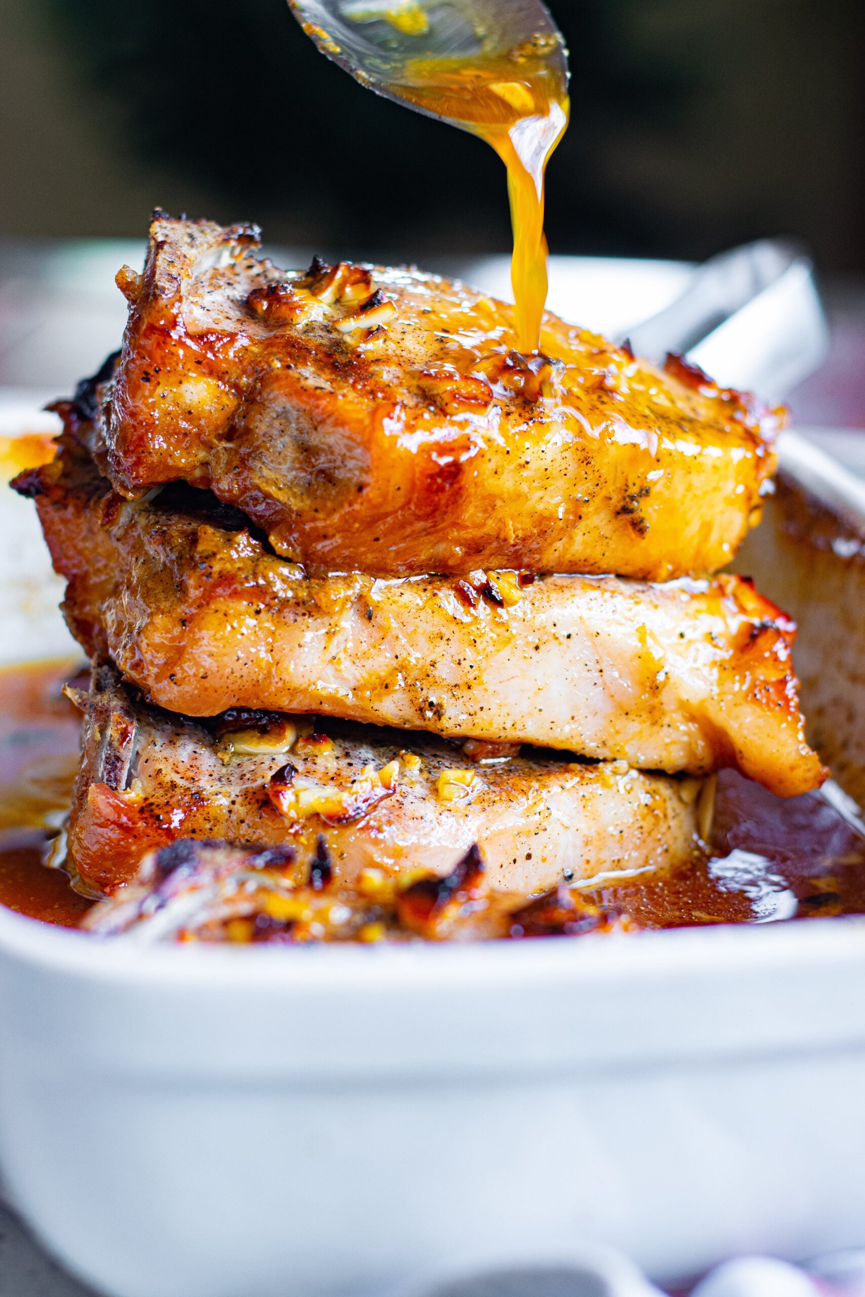 Front view of Honey Garlic Pork Chops stacked in a white baking dish with a silver spoon pouring honey garlic sauce over top.