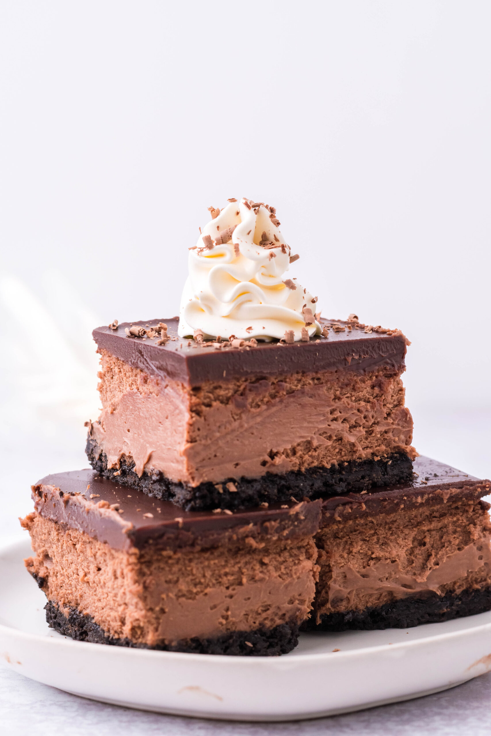 Three Chocolate Cheesecake bars topped on top of each other topped with whipped cream and chocolate shavings. 
