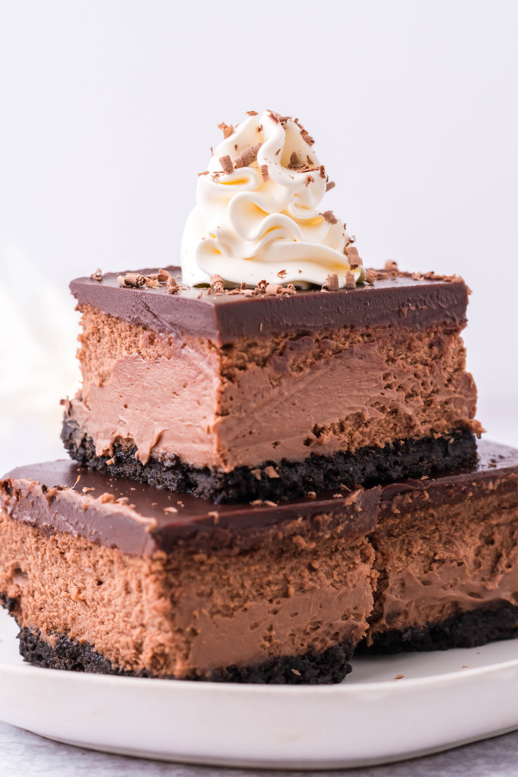 Three Chocolate Cheesecake bars topped on top of each other topped with whipped cream and chocolate shavings. 