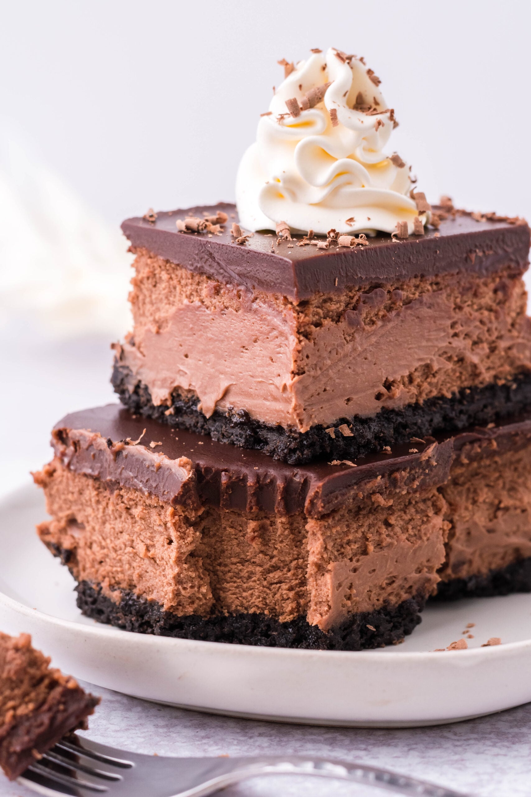Two Chocolate Cheesecake bars topped on top of each other topped with whipped cream and chocolate shavings. 