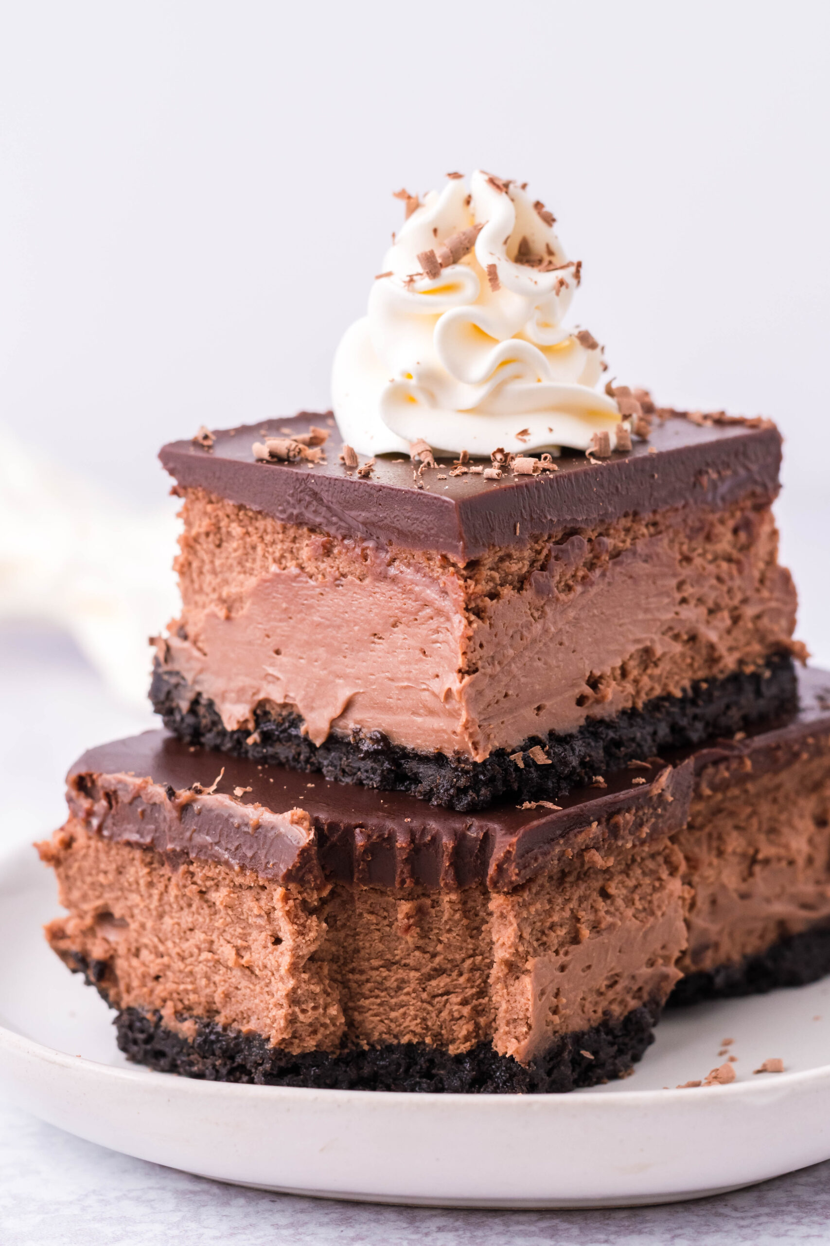 Two Chocolate Cheesecake bars topped on top of each other topped with whipped cream and chocolate shavings. 