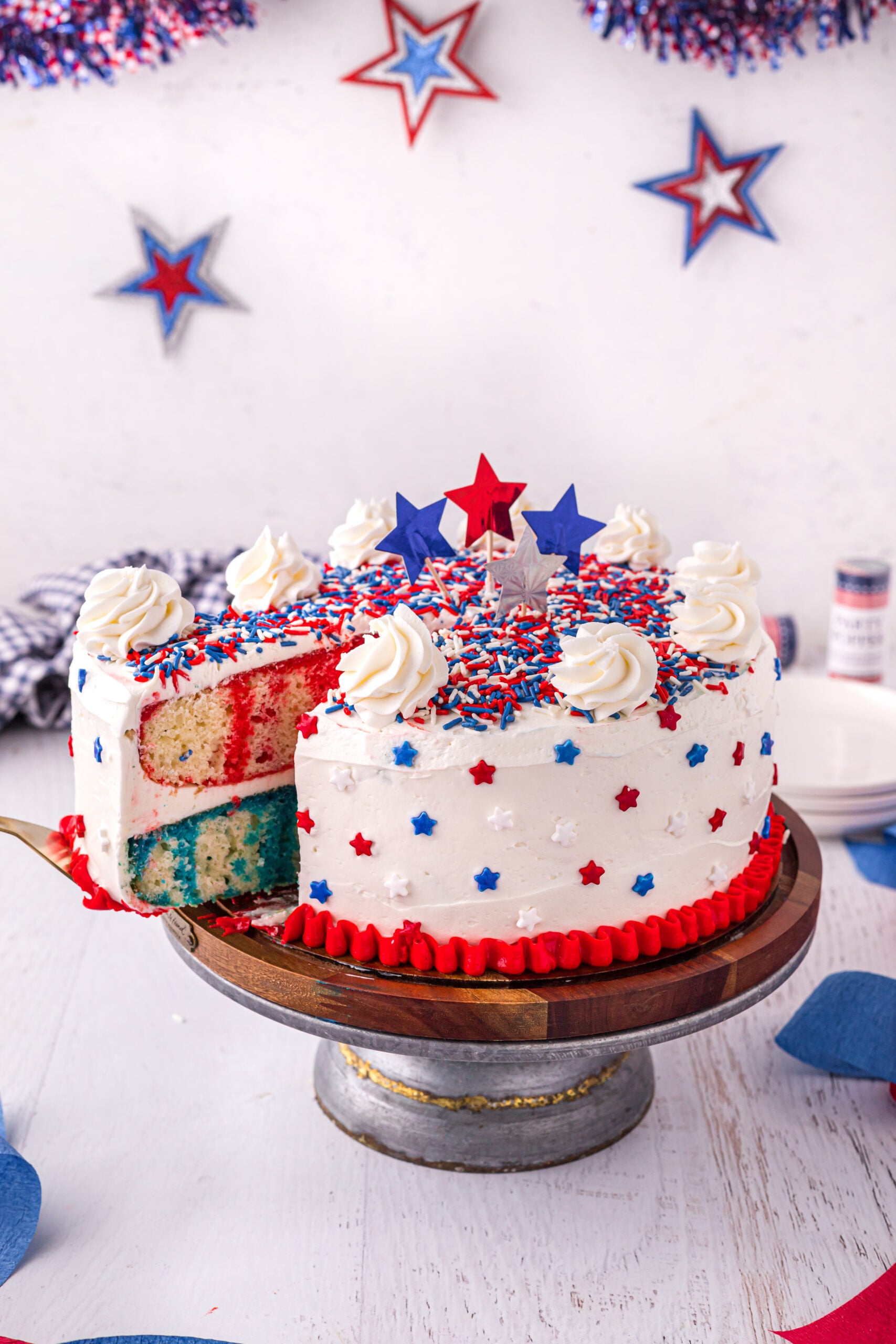Front view of 4th of July Cake on a wood cake stand with a slice being removed by a gold cake server.