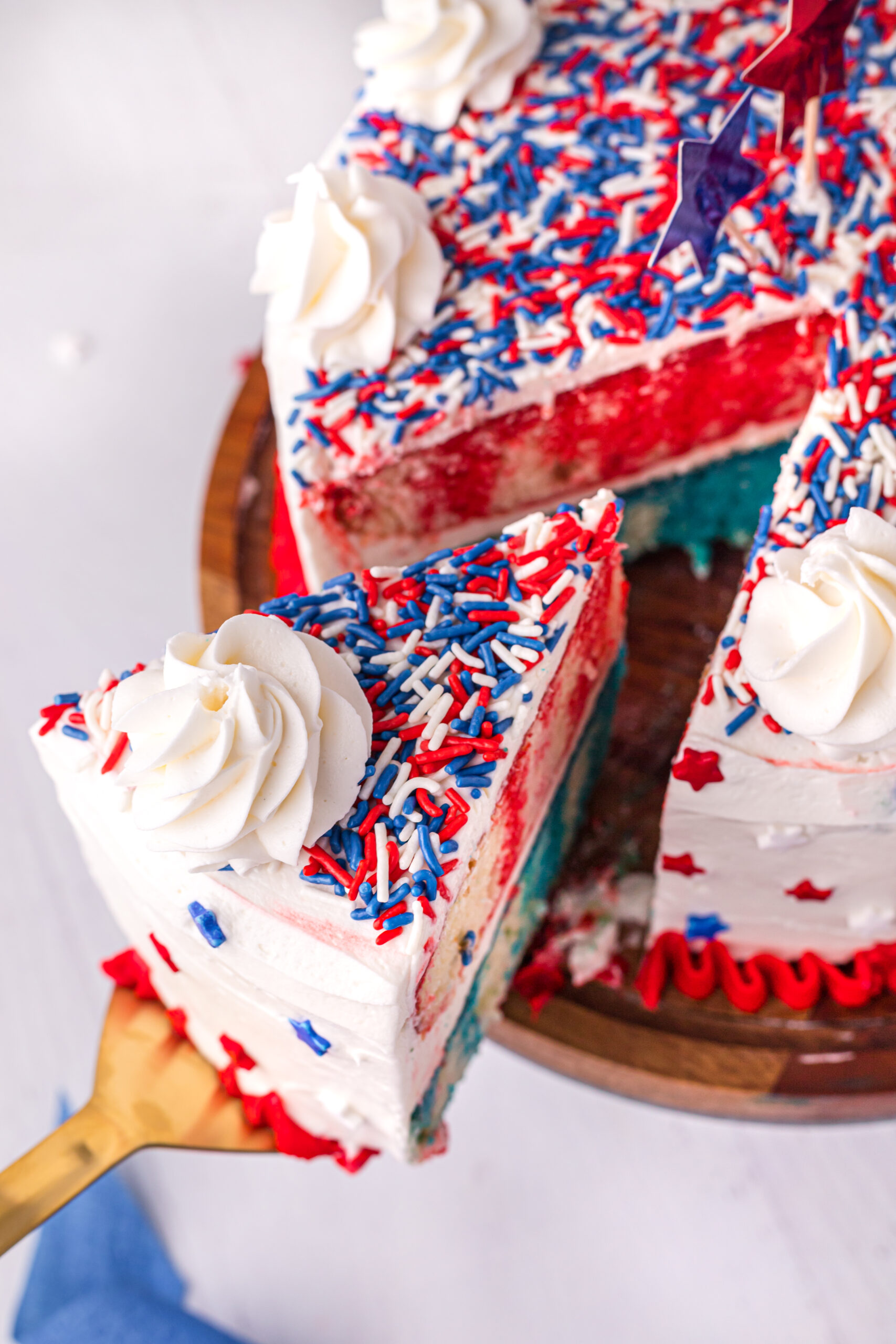 Close up view of 4th of July Cake on a wood cake stand with a slice being removed by a gold cake server.